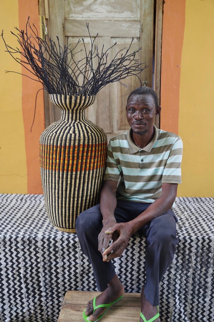 A man sitting next to a decorative woven piece with stylish branches on a black and white cloth against a wooden door.