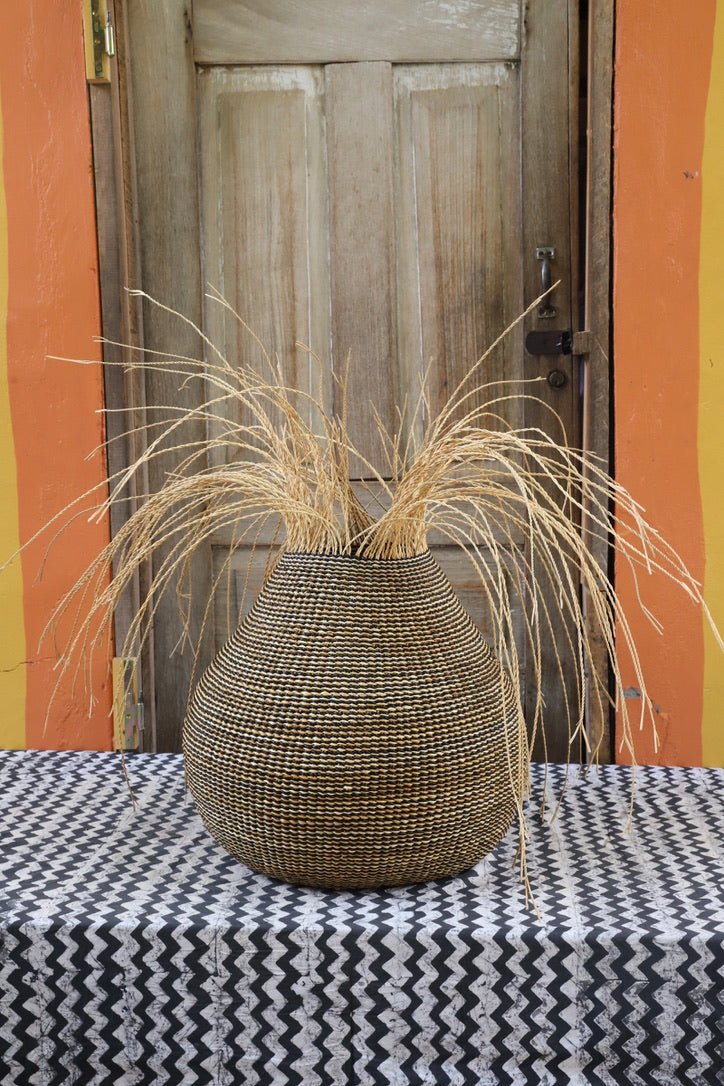 Patterned woven décor basket with straw branching out on a black and white cloth with a wooden door in the background.