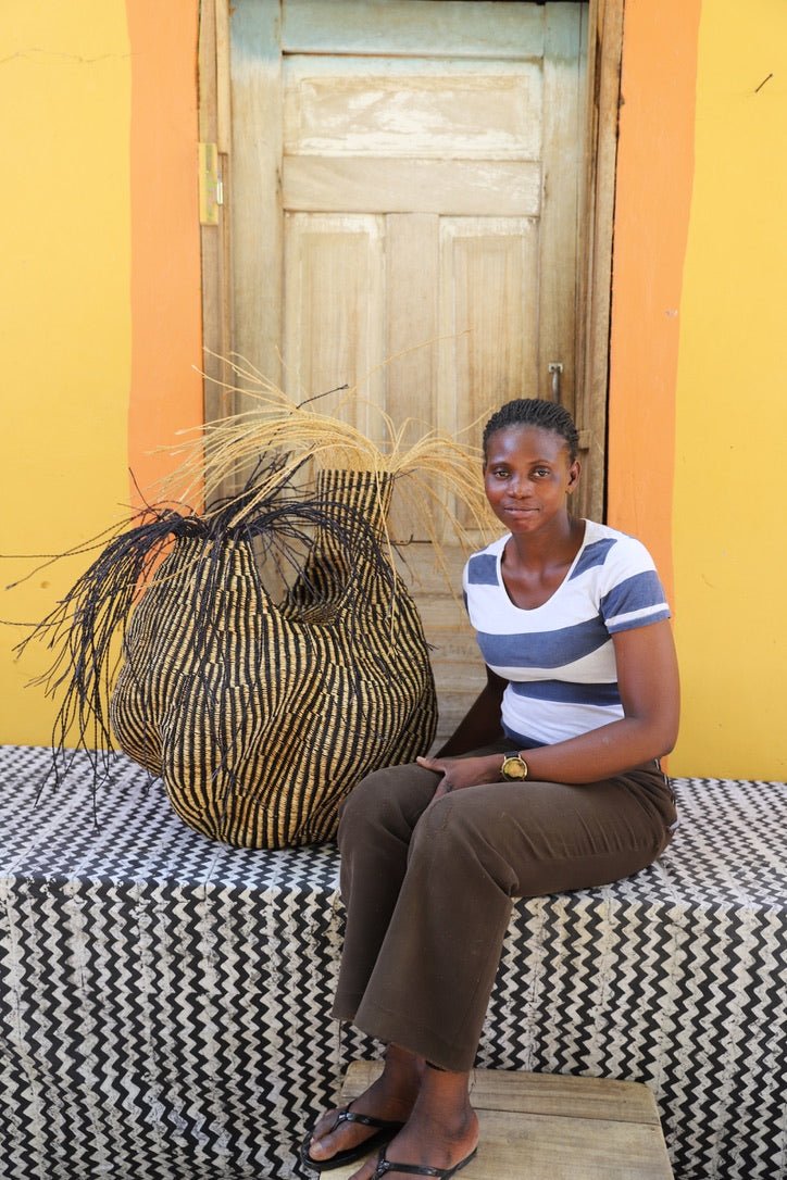 An artisan seated next to a handcrafted decorative basket with stylish straw branching out on a black and white cloth against a wooden door.