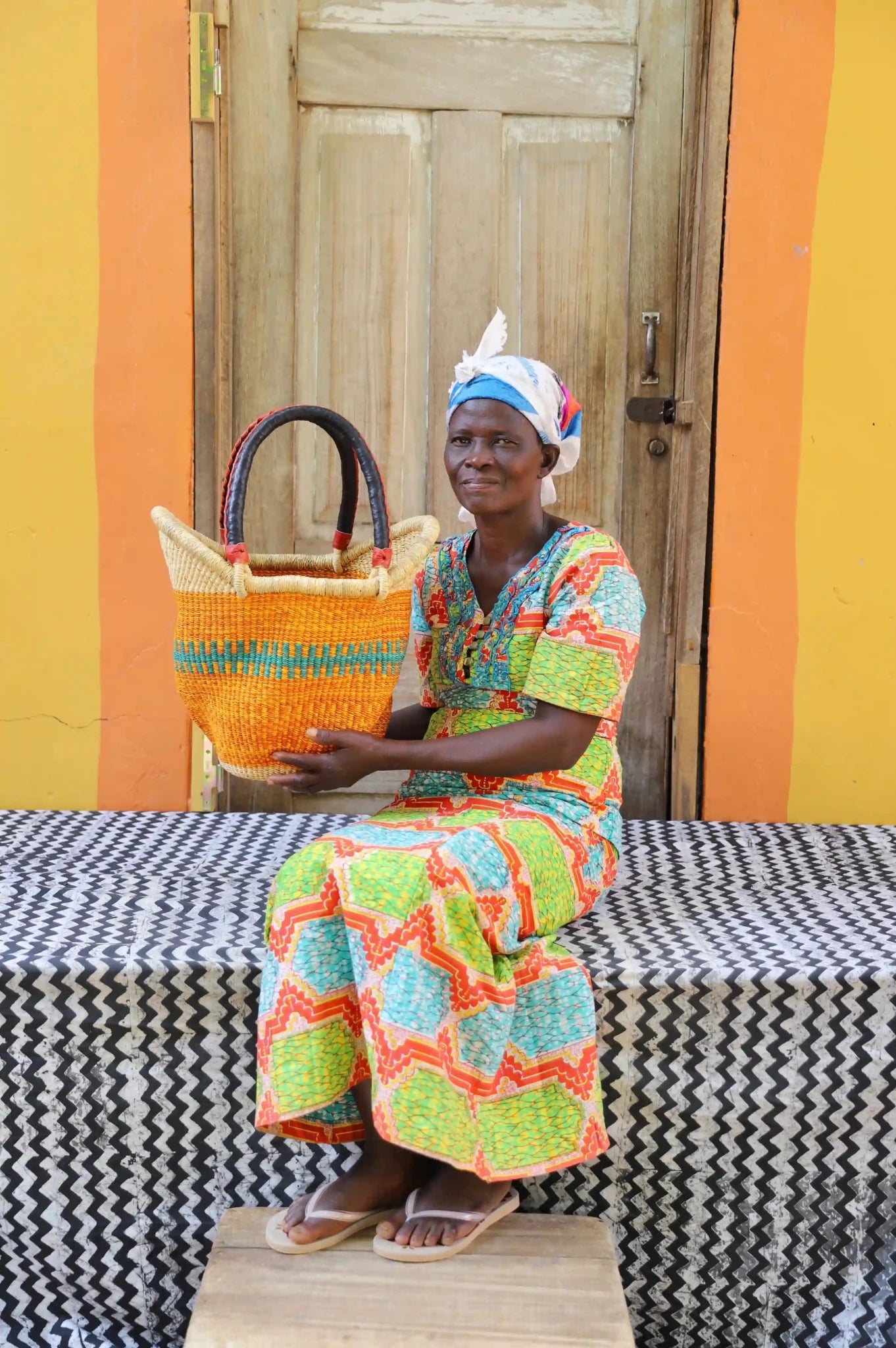 Woman in green and orange print dress holding a small amber Solara Carryall with teal accent stripe