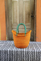 Orange woven basket with green handles on a tablecloth in front of a wooden door.