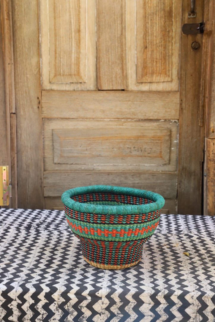 Woven basket with teal top and red and green pattern on a textured surface in front of a wooden door.