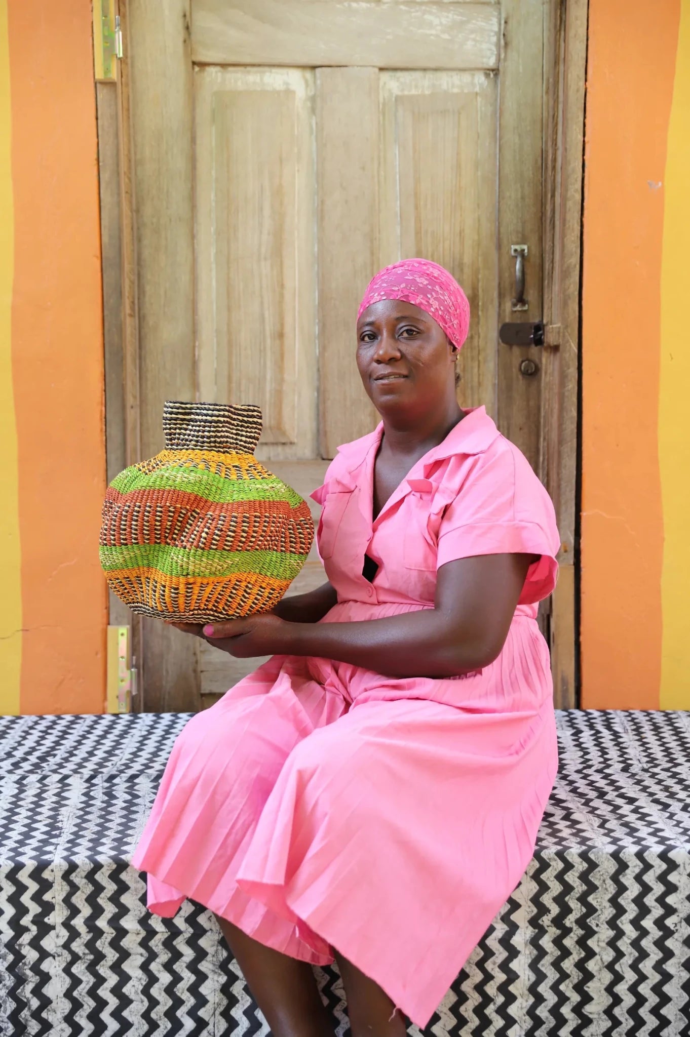 An artisan seated next to a handcrafted decorative basket  on a black and white cloth against a wooden door.