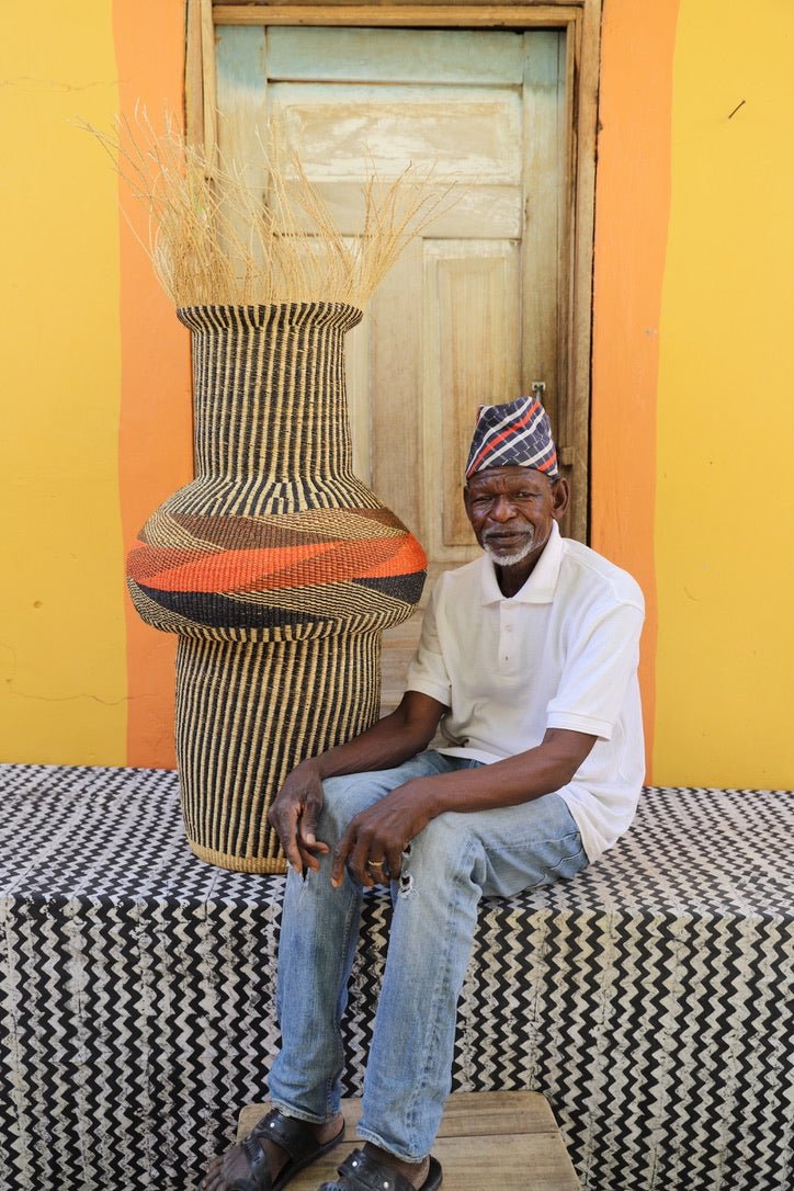 Man sitting next to a colorful woven basket with stylish branches on a black and white patterned surface with a wooden door in the background.