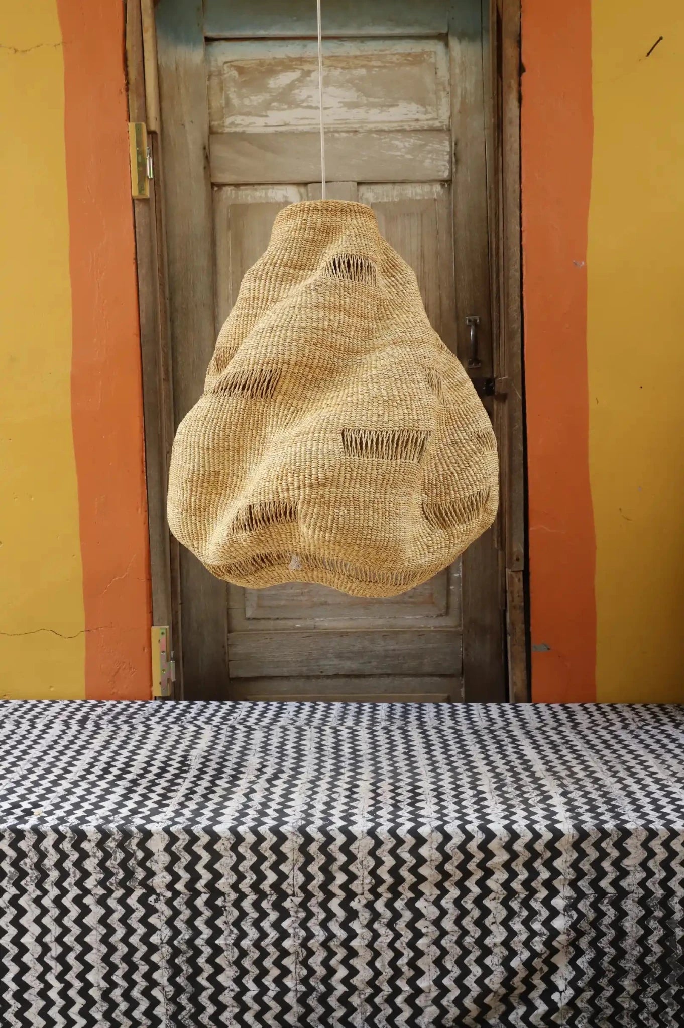Woven basket hanging from a string against a yellow wall with a wooden door.