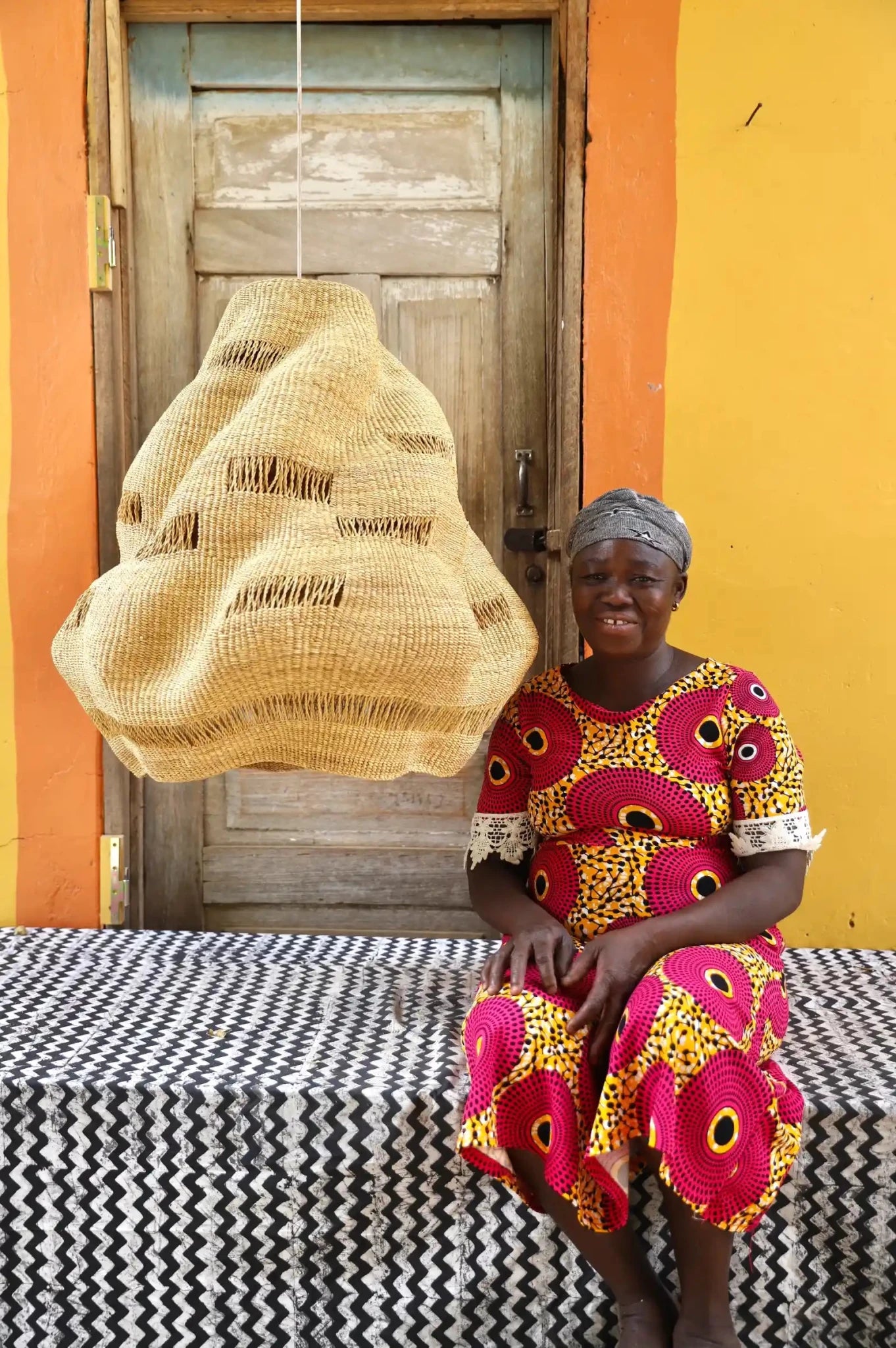 Woman in a colorful dress sitting on a bench with a woven basket behind her against a yellow wall.