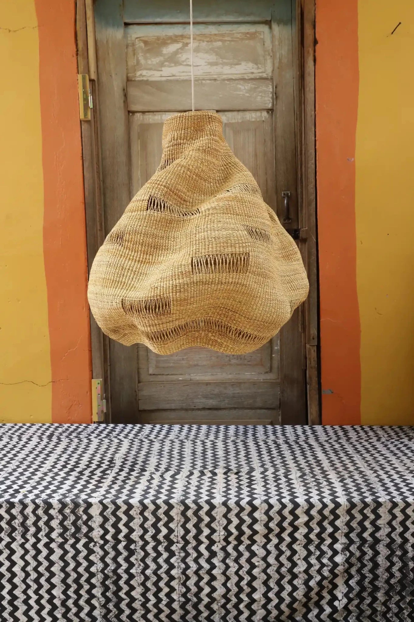 Woven basket hanging above a patterned tablecloth with a wooden door in the background.