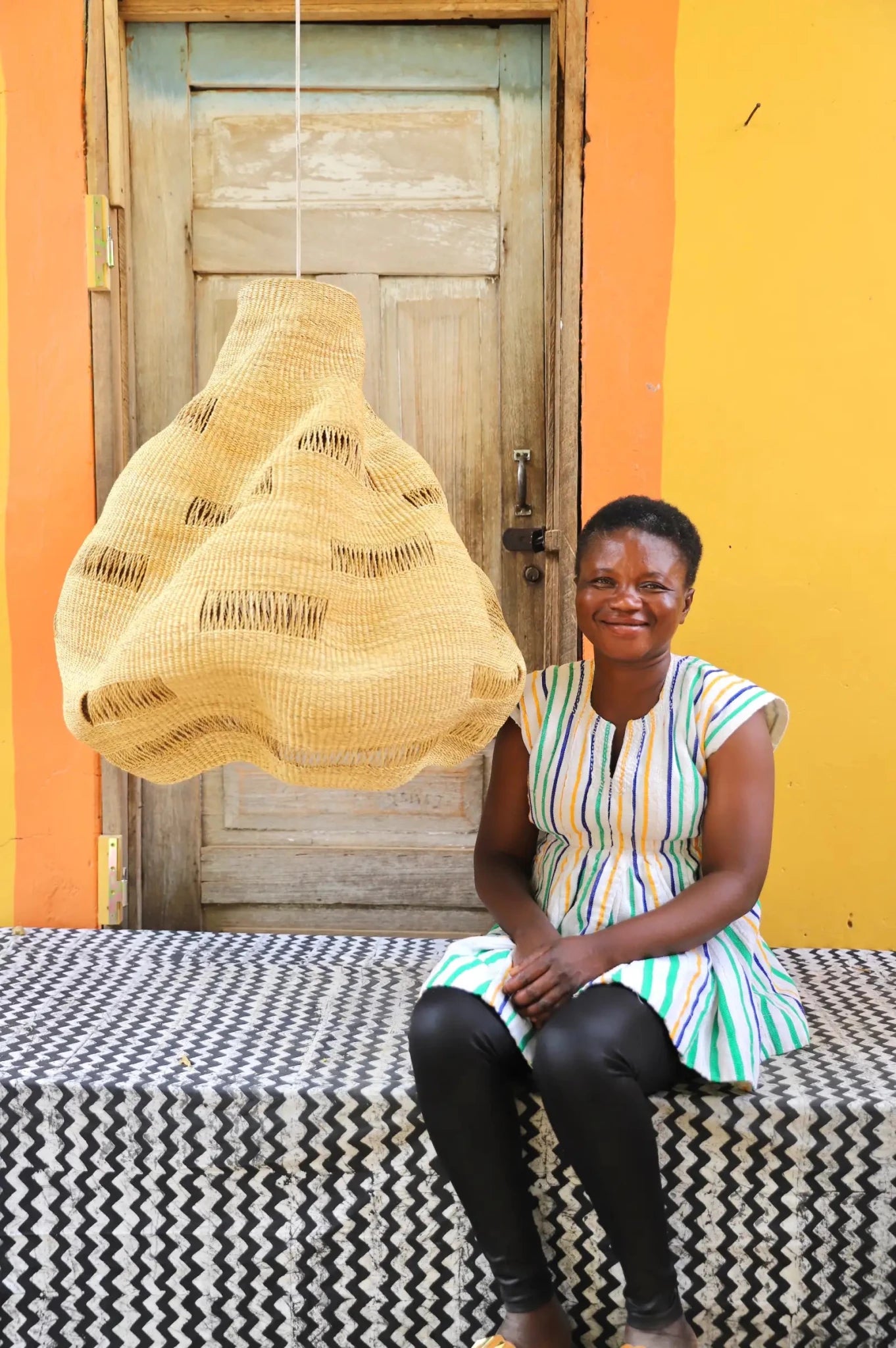 Woman sitting on a black and white cloth with a woven basket against a colorful wall.