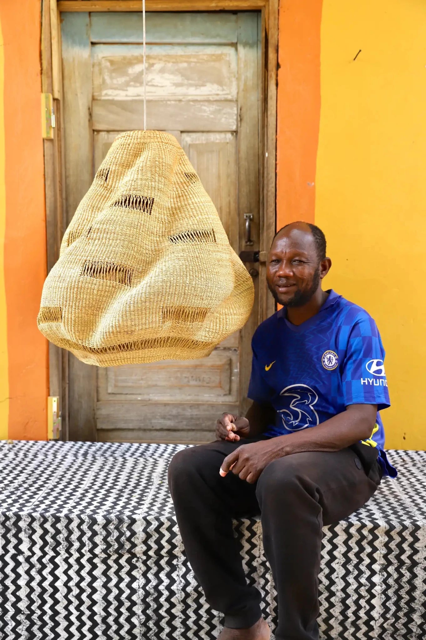Man sitting on a patterned bench with a woven basket above him against a colorful wall.