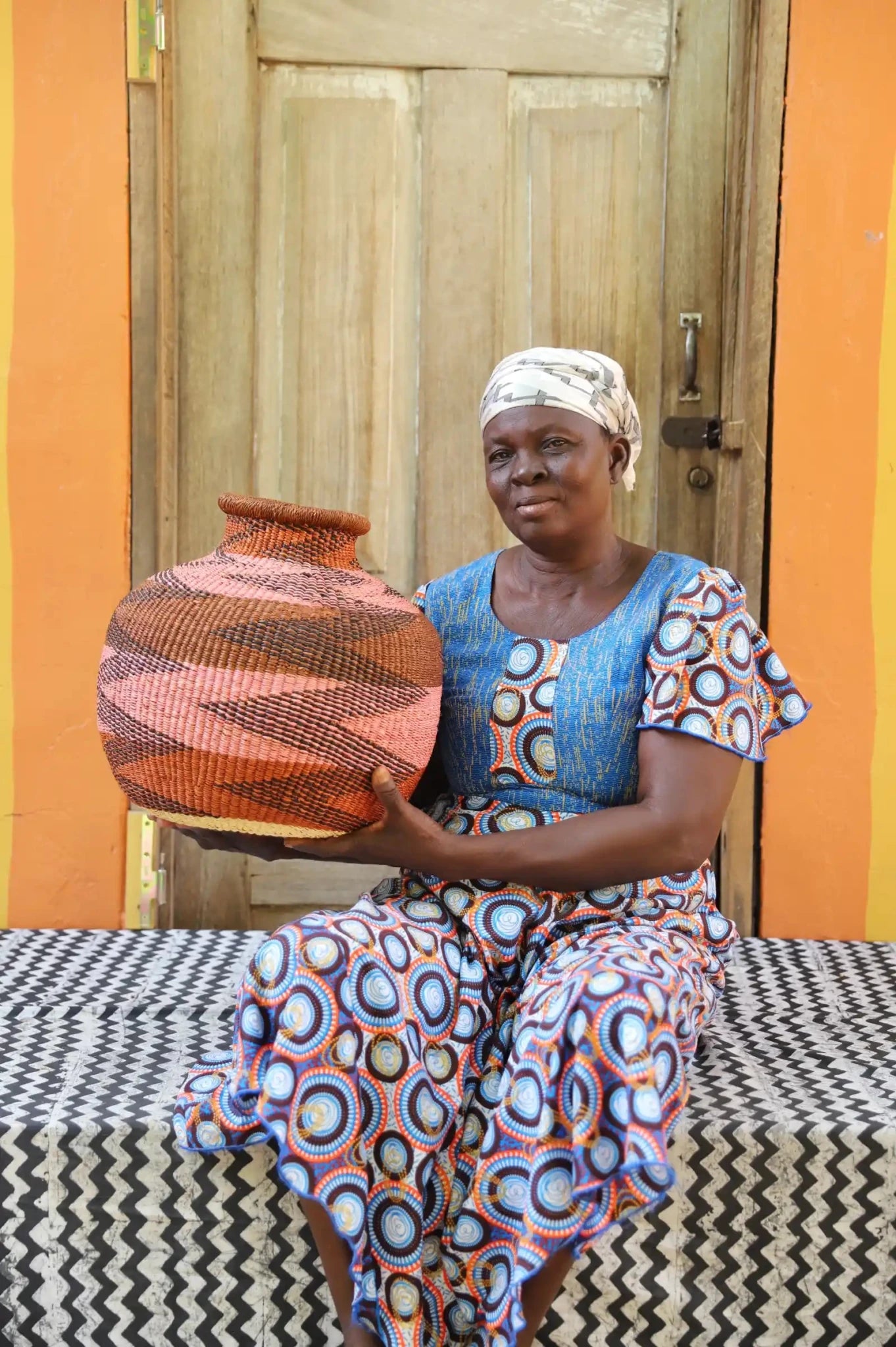 Artisan holding a colorful woven storage basket  on a black and white cloth against a wooden door.
