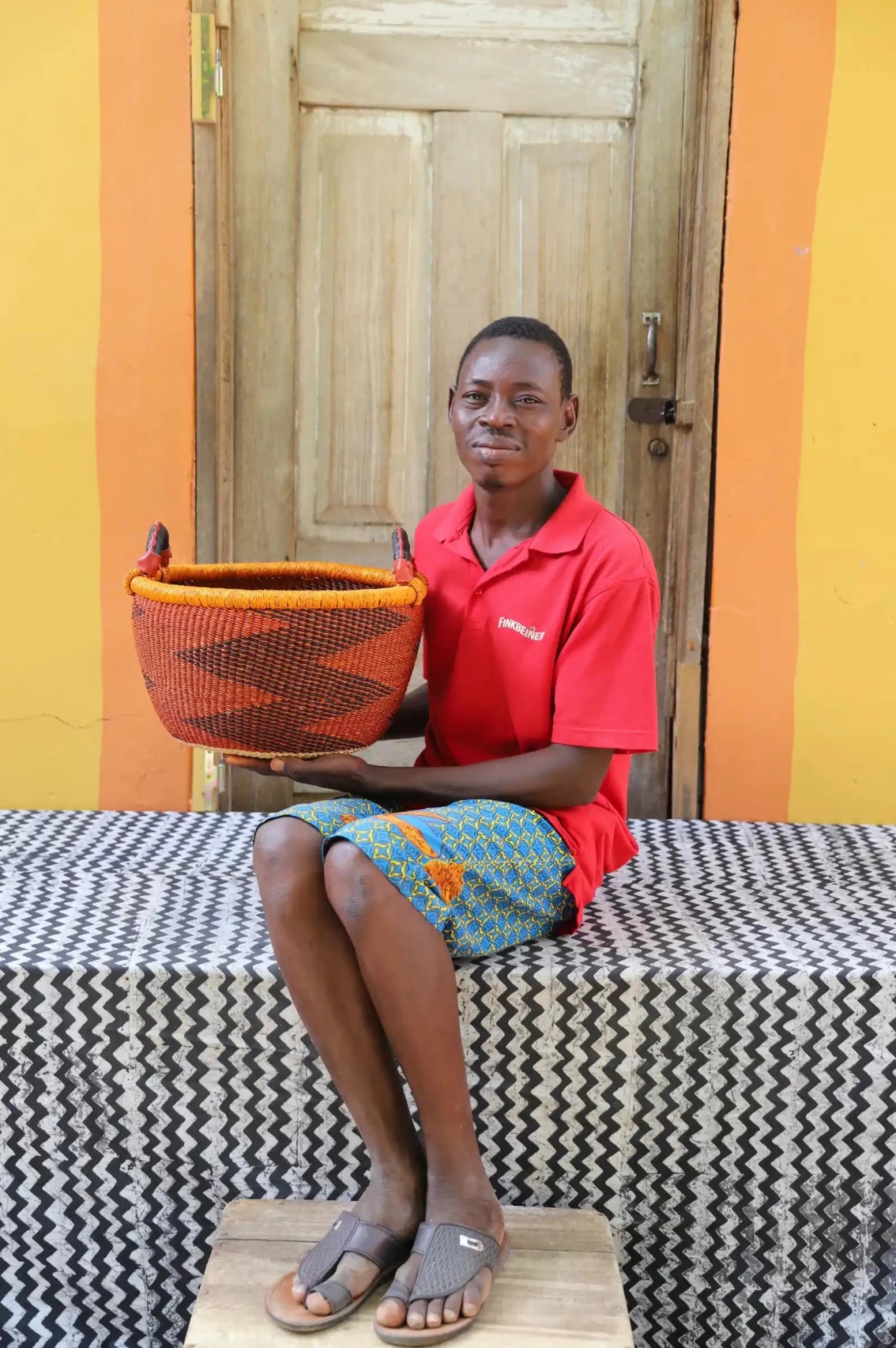 Artisan holding a colorful handwoven storage basket sitting on a black and white cloth against a wooden door.