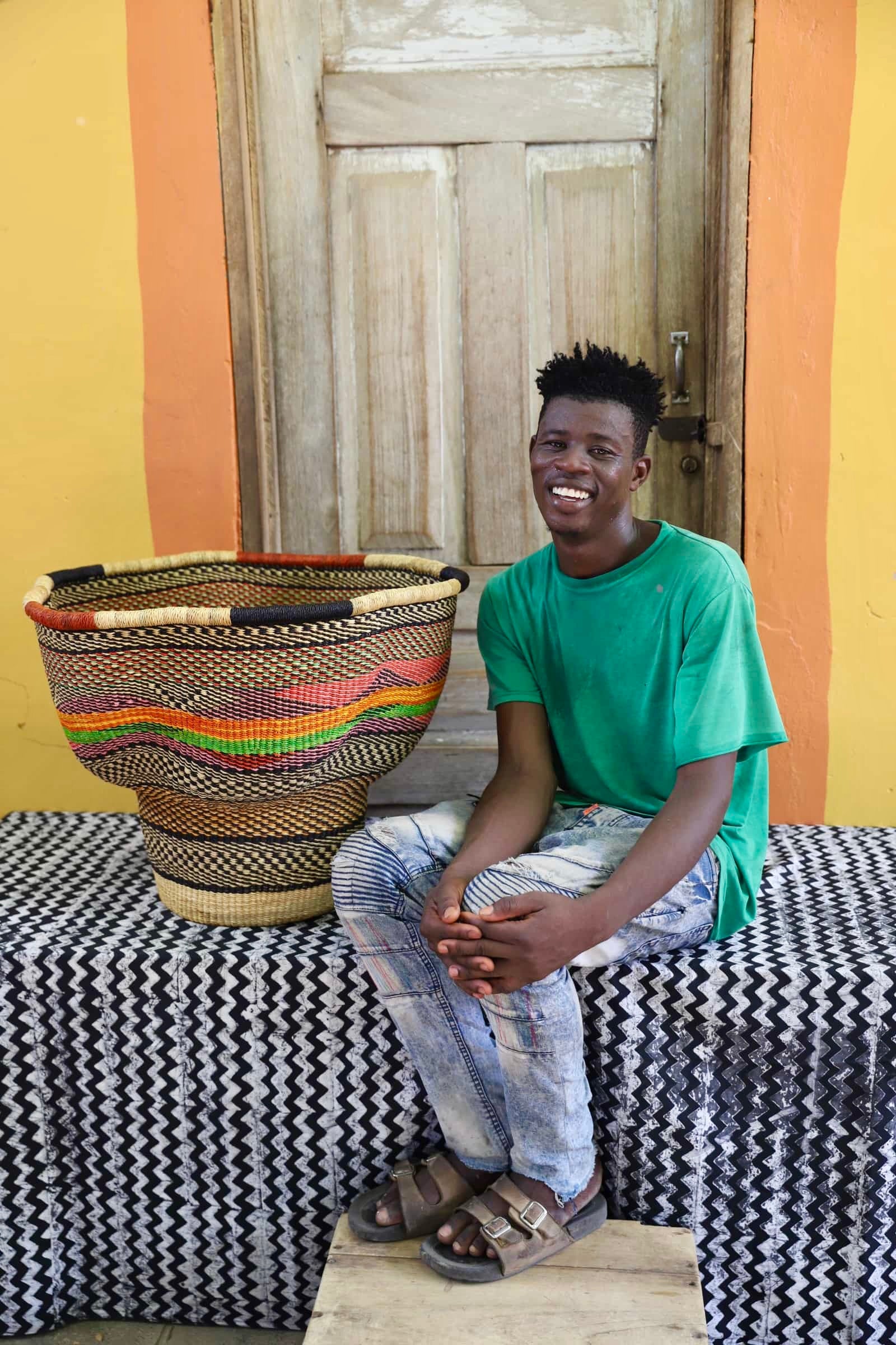 An artisan seated next to a handcrafted decorative basket with stylish straw branching out on a black and white cloth against a wooden door.