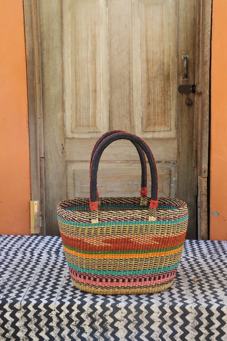 Round carry basket with teal, terracotta and orange horizontal stripe bands, dark leather handles, against a wooden door.