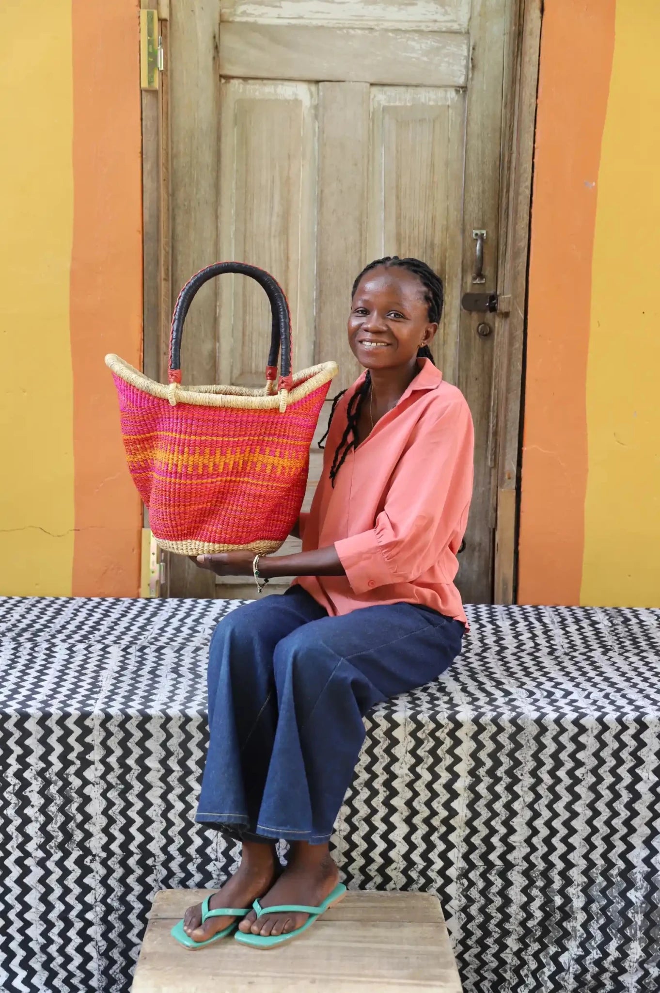 Woman in peach shirt seated holding a medium Solara Carryall with vibrant pink and orange stripe