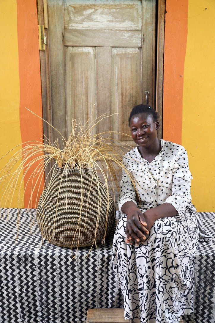 Woman sitting next to a patterned woven décor basket with straw branching out on a black and white cloth with a wooden door in the background.
