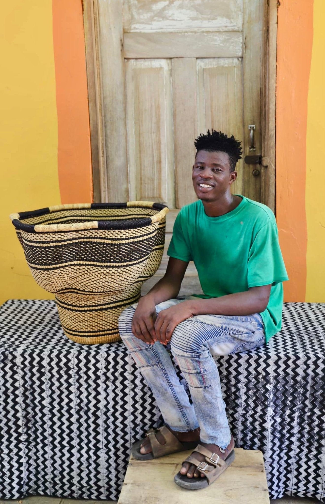 An artisan seated next to a handcrafted decorative basket with stylish straw branching out on a black and white cloth against a wooden door.