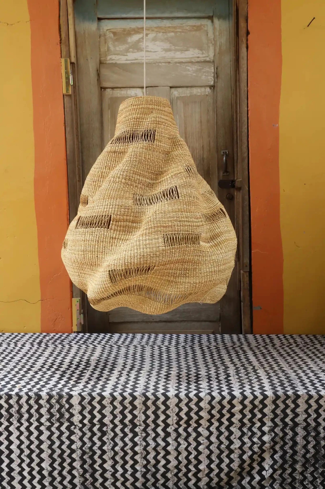 Handwoven Cosma pendant lampshade in natural straw, with a teardrop open-weave form, hanging before a wooden door.