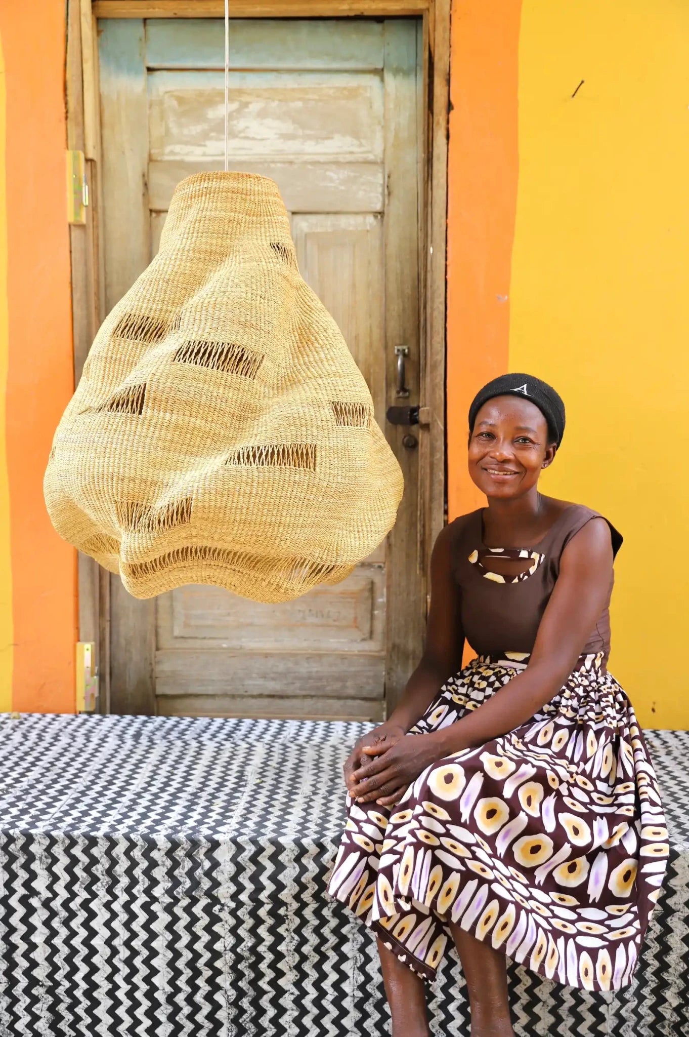 Woman seated beside a handwoven Cosma pendant lampshade in natural straw with a teardrop open weave, before a wooden door.