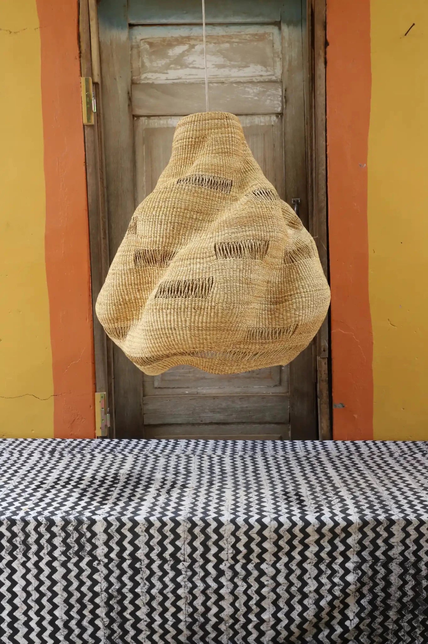 Handwoven Cosma pendant lampshade in natural straw with an open weave and organic form, hanging before a wooden door.