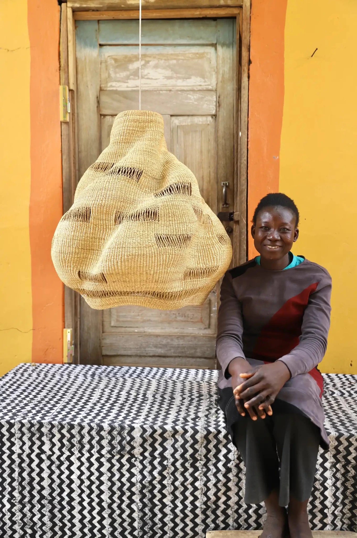 Person seated beside a handwoven Cosma pendant lampshade in natural straw with an open weave, before a wooden door.