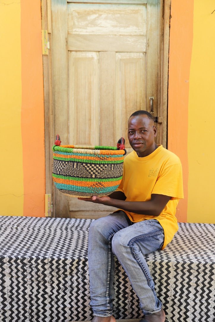 Artisan holding a colorful handwoven storage basket sitting on a black and white cloth against a wooden door.