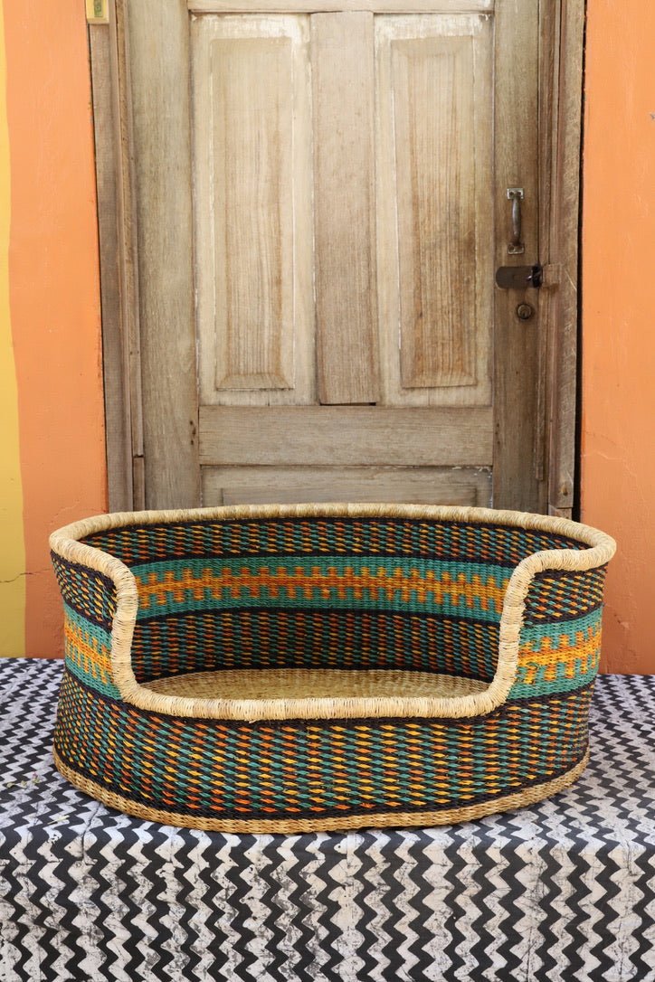 Woven pet bed with colorful patterns on a patterned rug in front of a wooden door.
