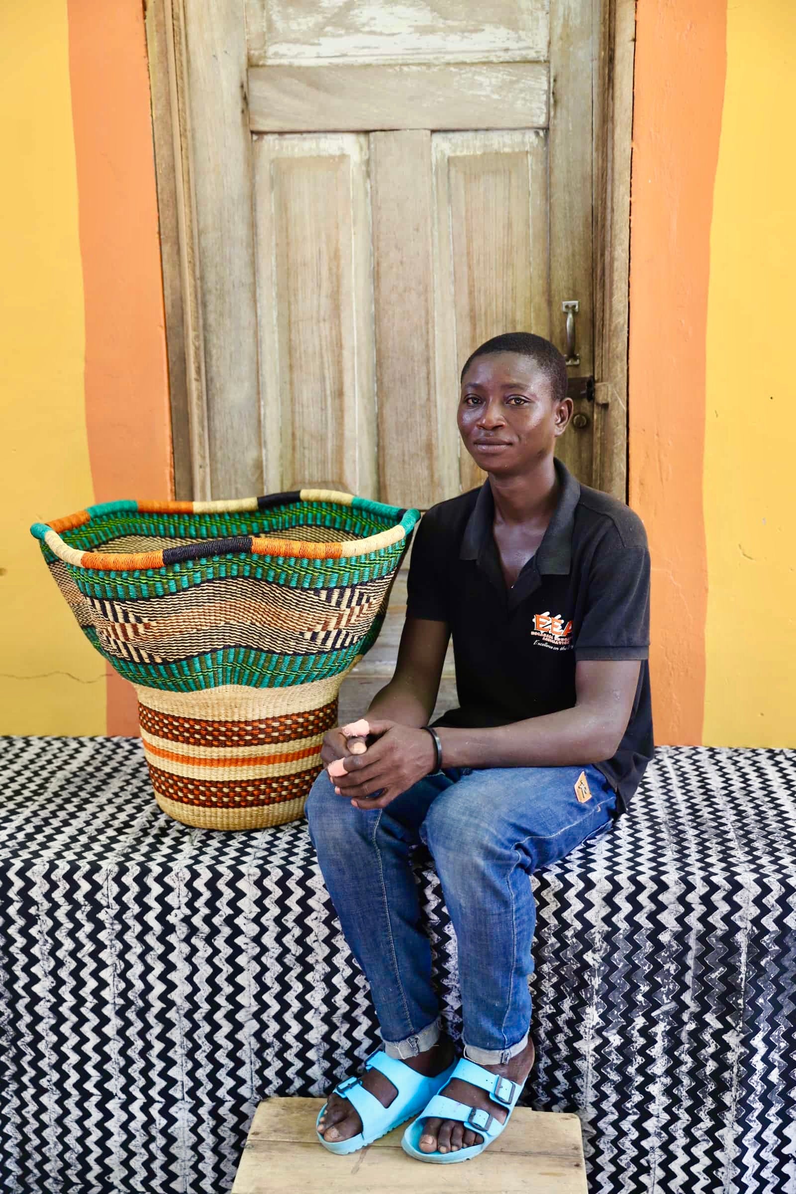 An artisan seated next to a handcrafted decorative basket with stylish straw branching out on a black and white cloth against a wooden door.
