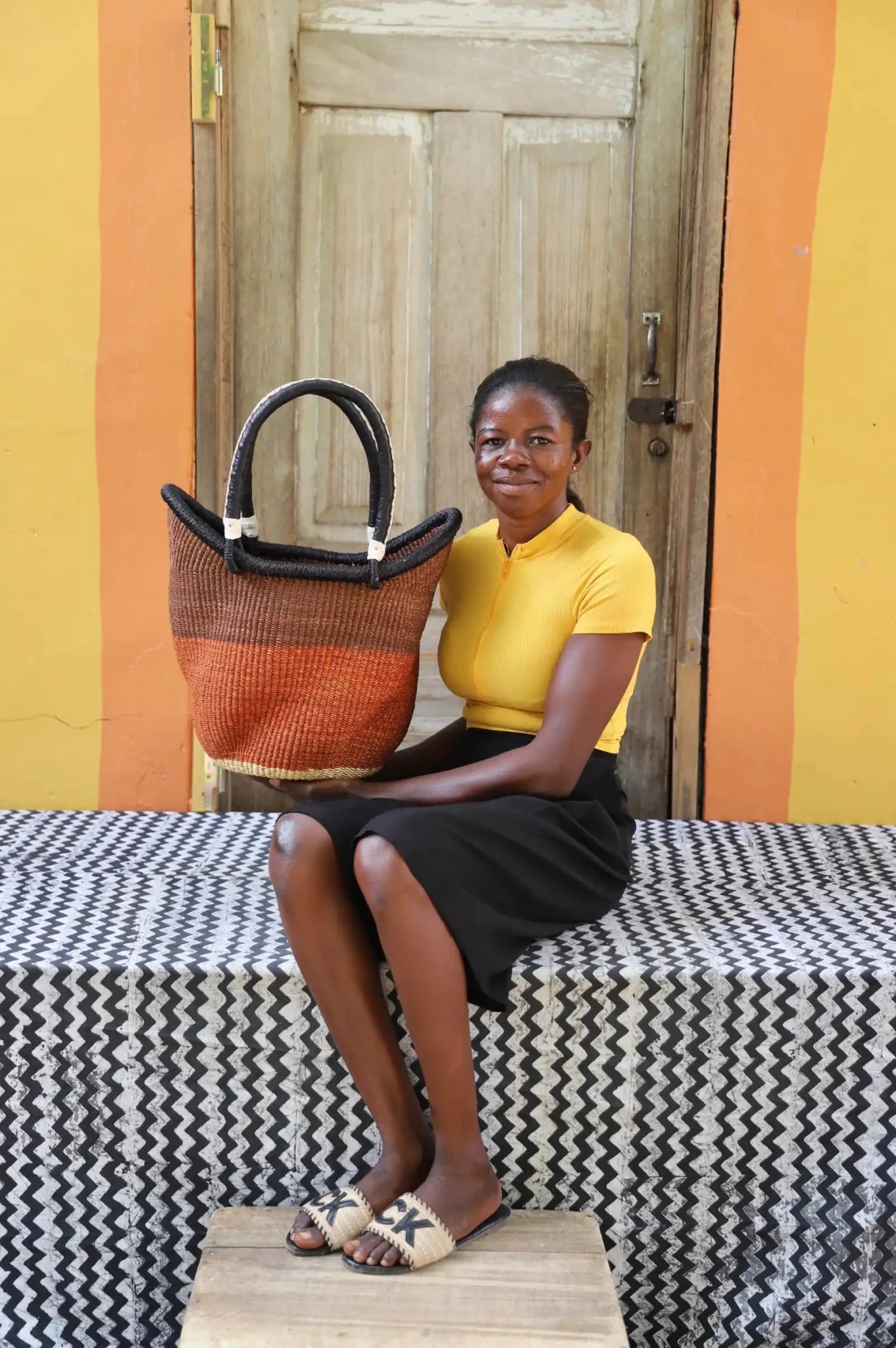 Woman in yellow top and black skirt holding a medium Solara Carryall with rust and tan colour block