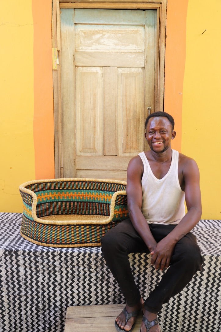 Man sitting on a patterned bench with a colorful woven Dog Bed next to him against a yellow wall.