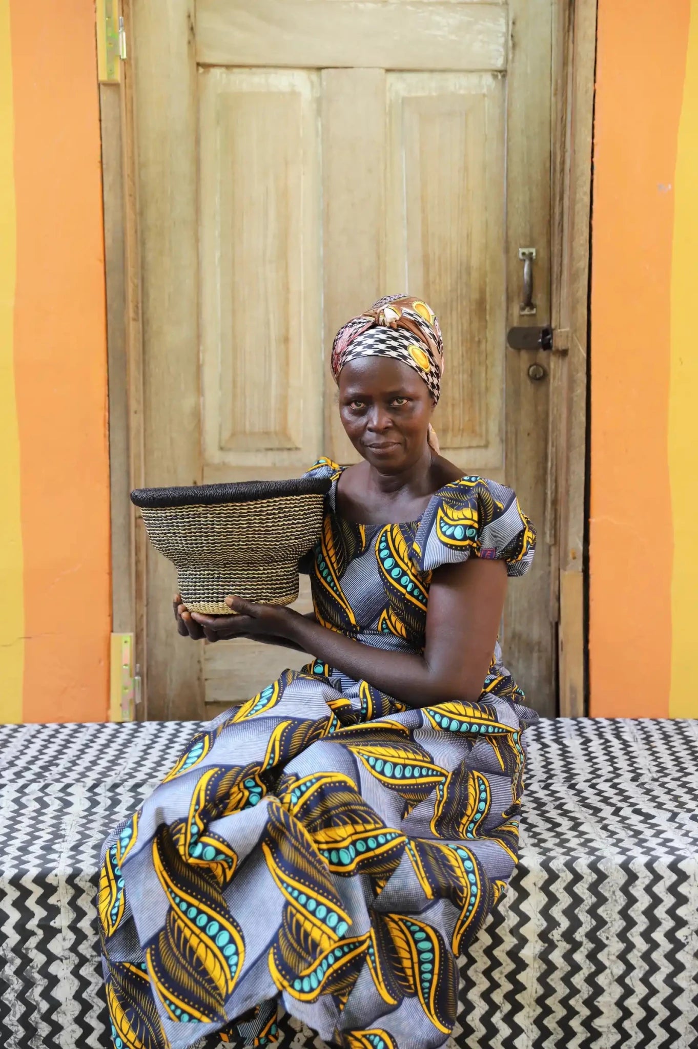 Woman in a patterned dress holding a basket in front of a wooden door with an orange wall.