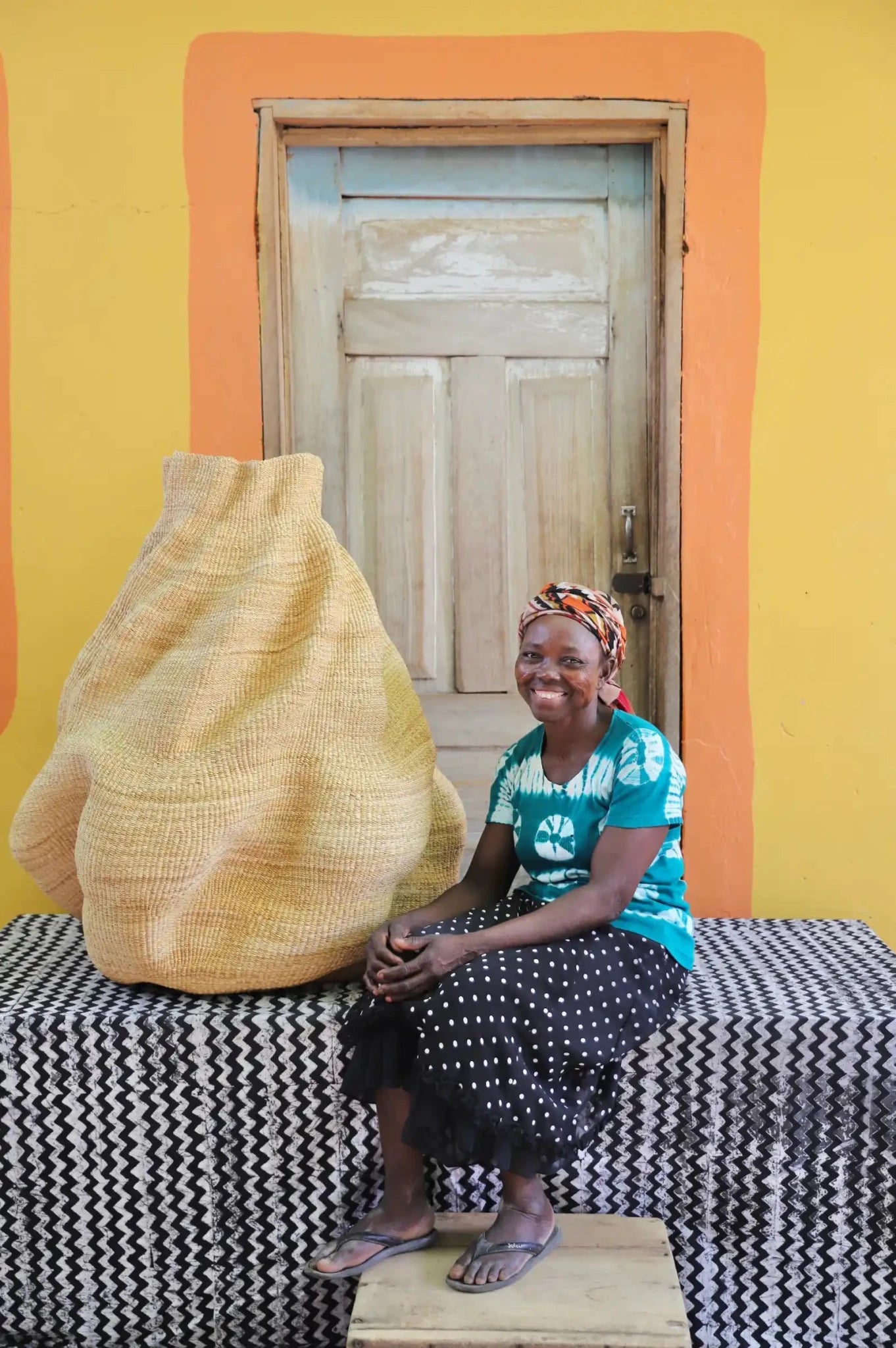 An artisan seated next to a handcrafted decorative basket  on a black and white cloth against a wooden door.