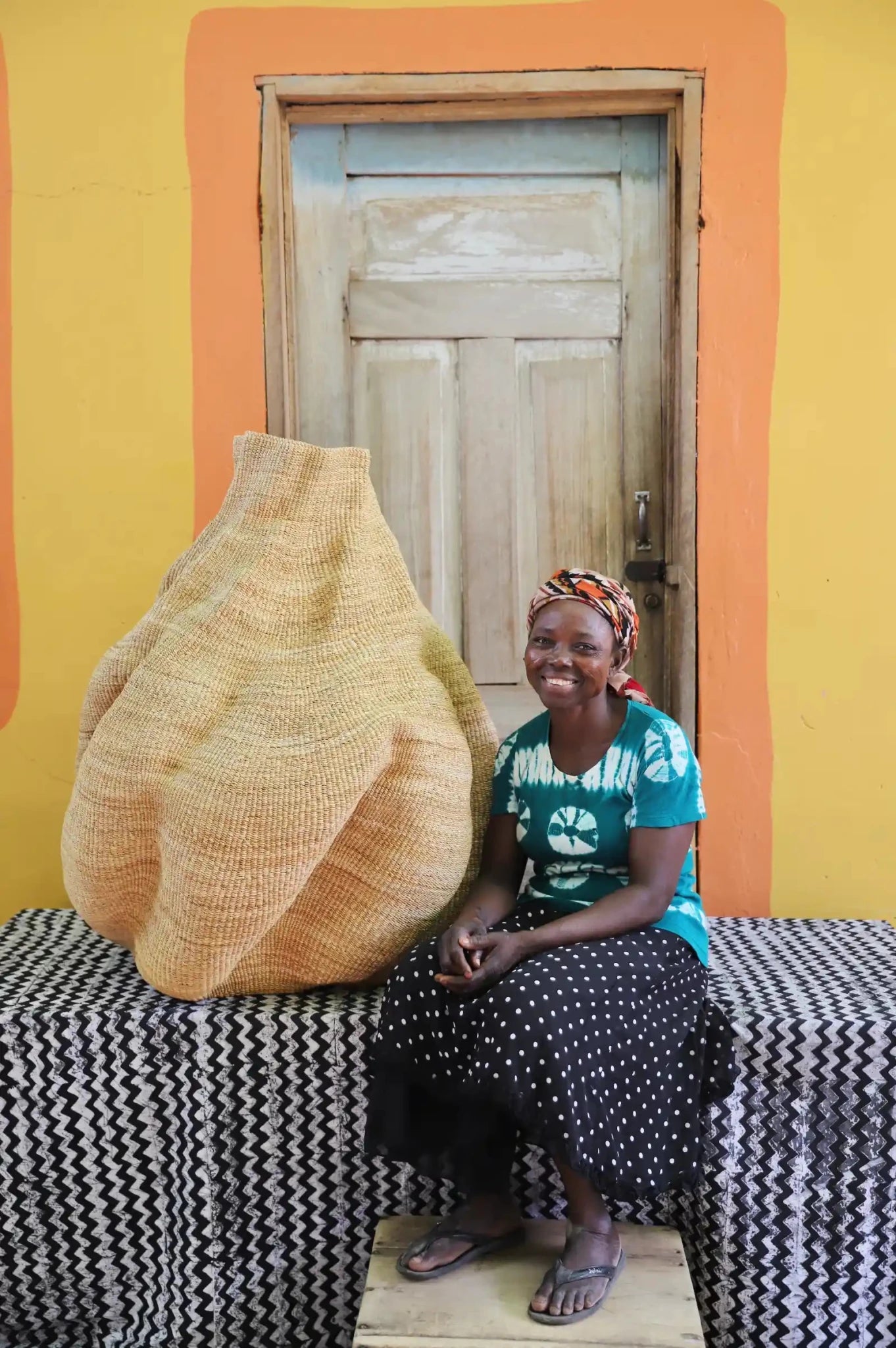 An artisan seated next to a handcrafted decorative basket  on a black and white cloth against a wooden door.