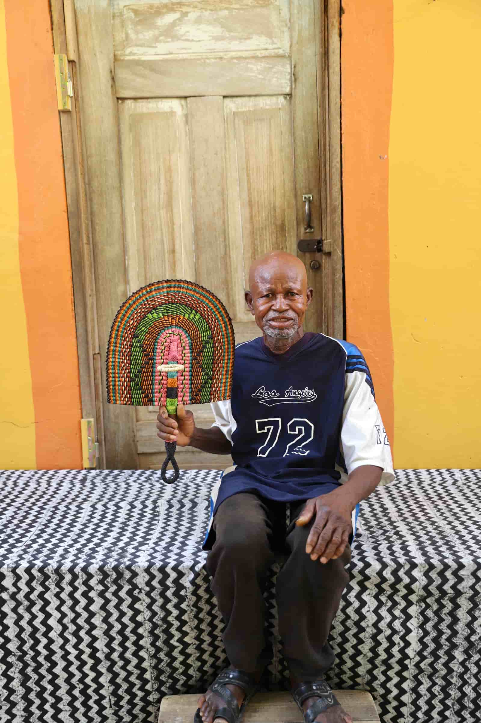 Man sitting on a black and white cloth  holding a decorative fan in front of a colorful wall.