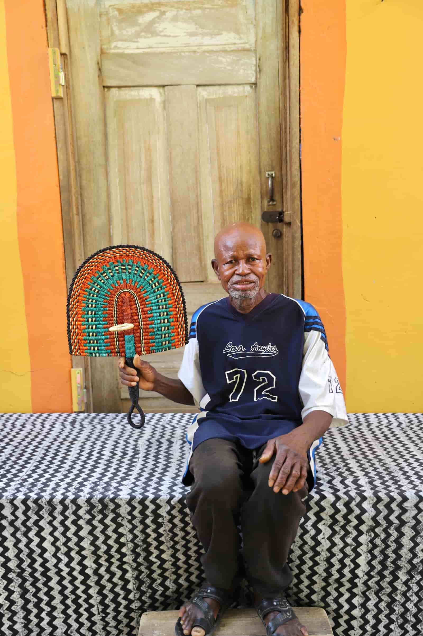 Man sitting on a bench holding a colorful fan against a colorful wall.