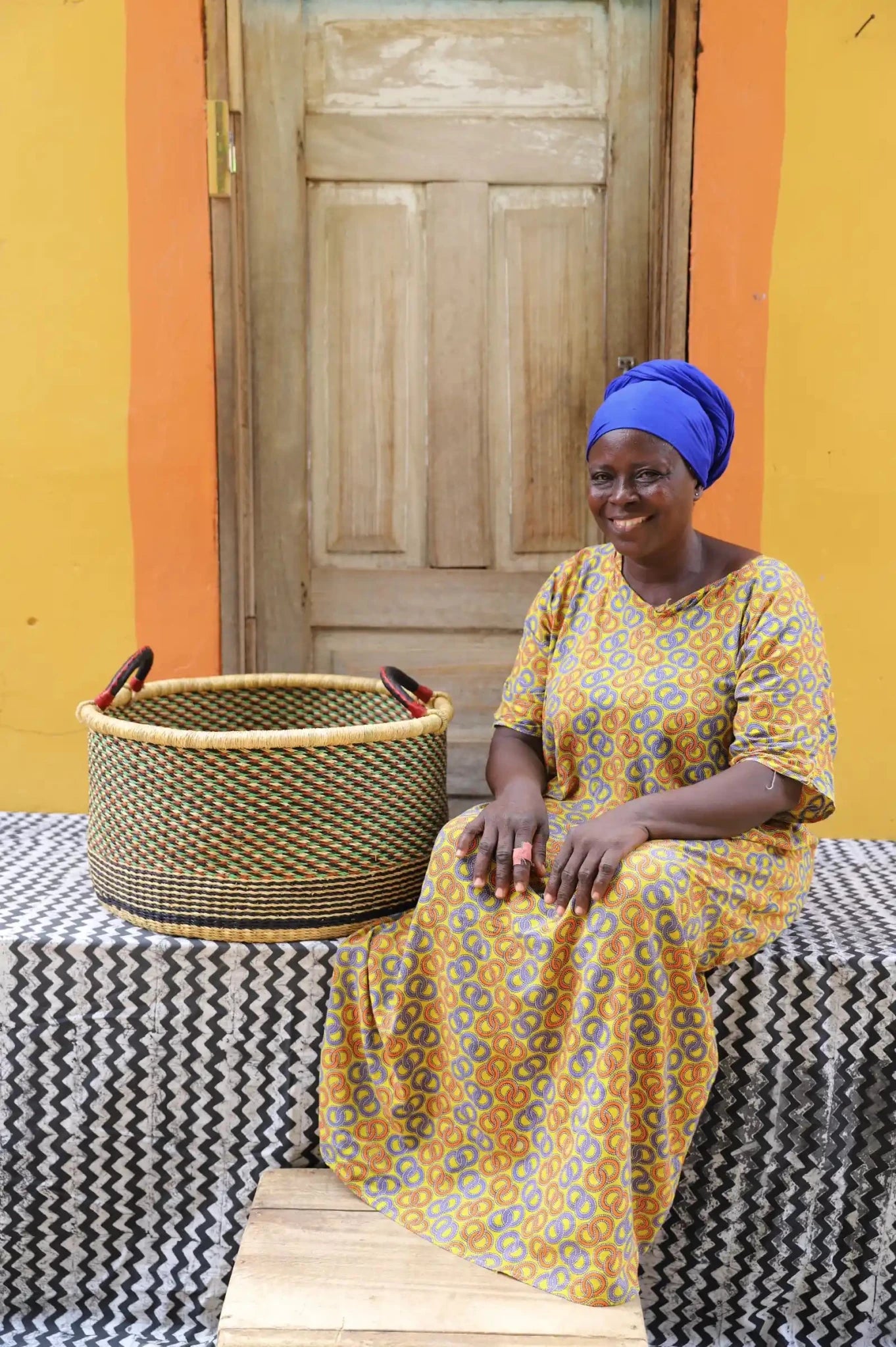Woven Tub (Large) with woman weaver handwoven in Ghana sitting on black and white cloth and wooden door in background

