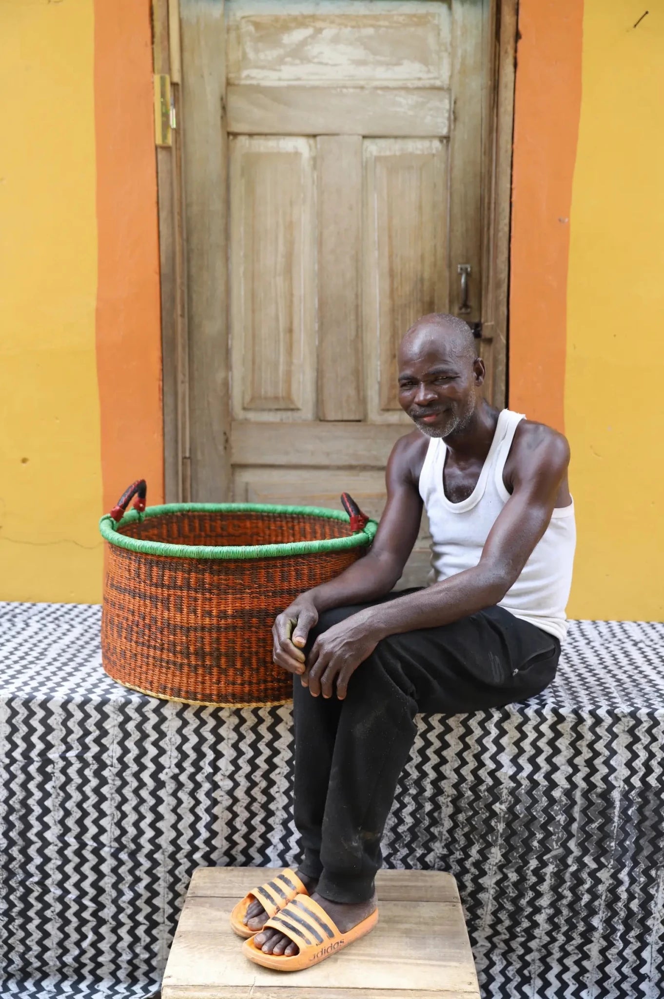 Woven Tub (Large) with man weaver handwoven in Ghana sitting on black and white cloth and wooden door in background

