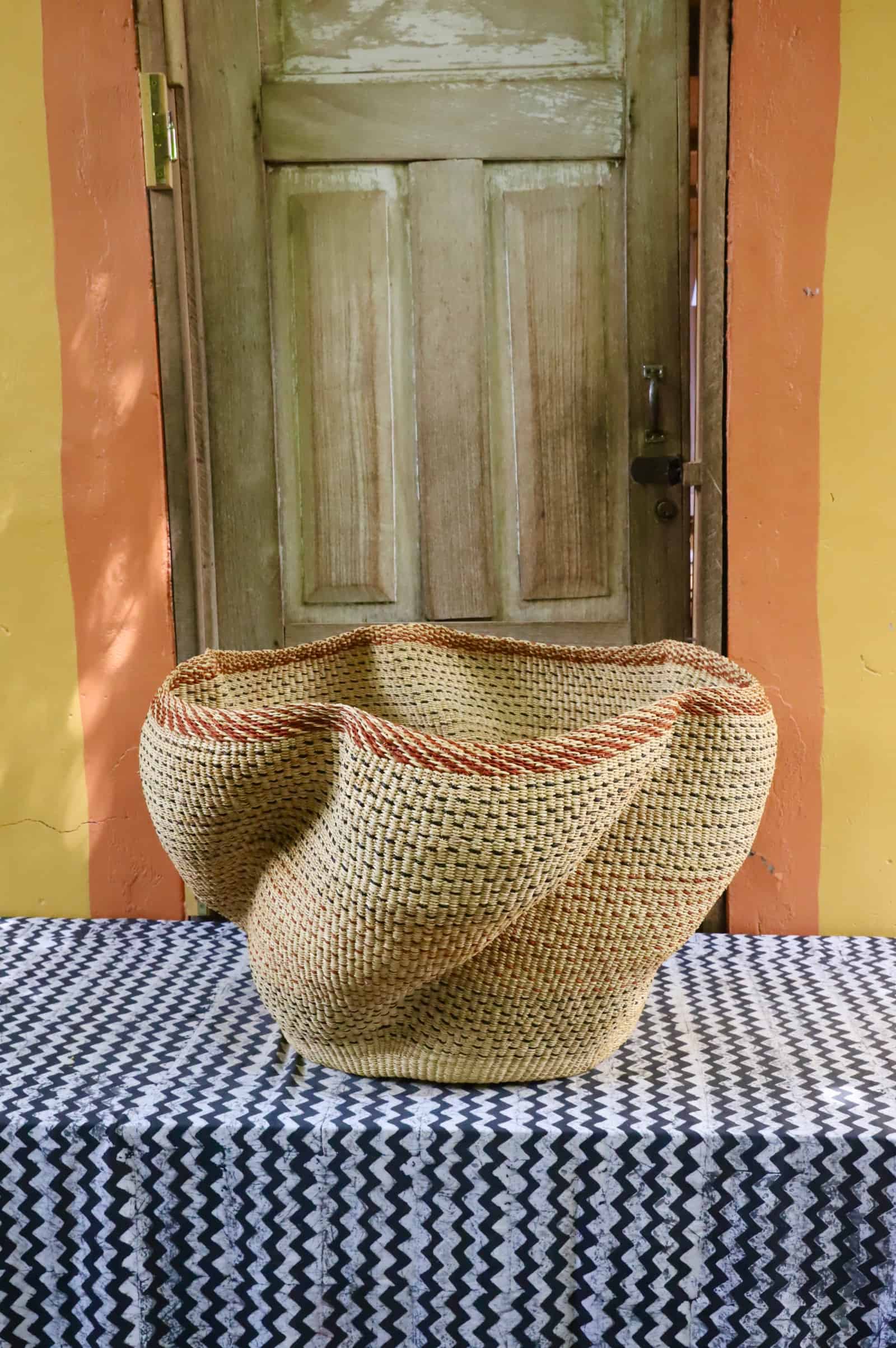 Giant Wave art basket in natural straw with a terracotta rim stripe, on a black and white cloth before a wooden door.