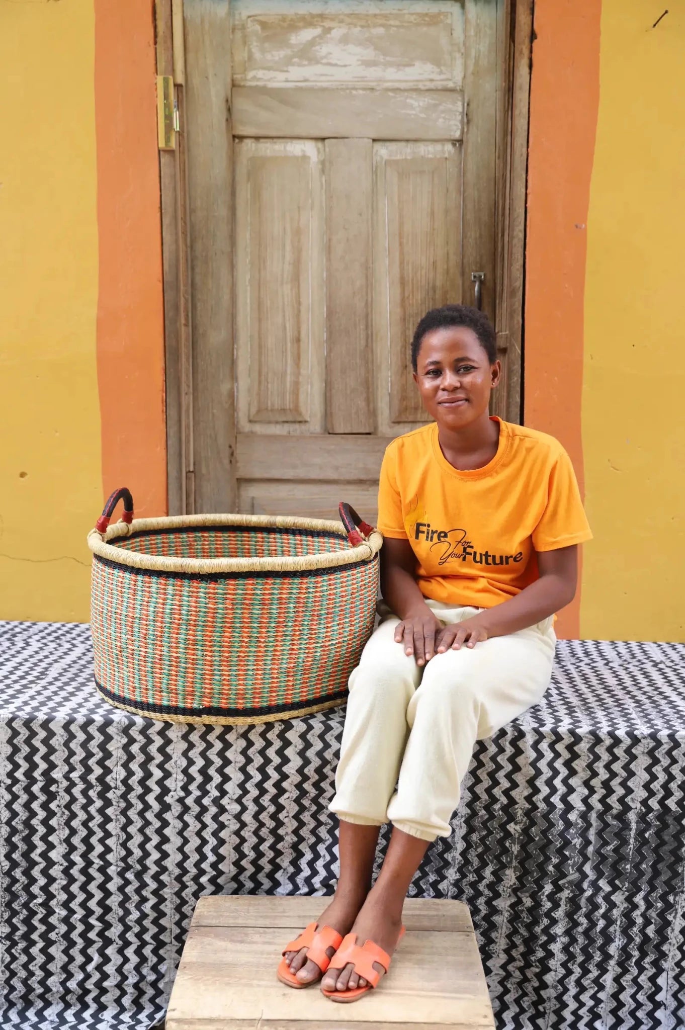 Woven Tub (Large) with woman weaver handwoven in Ghana sitting on black and white cloth and wooden door in background