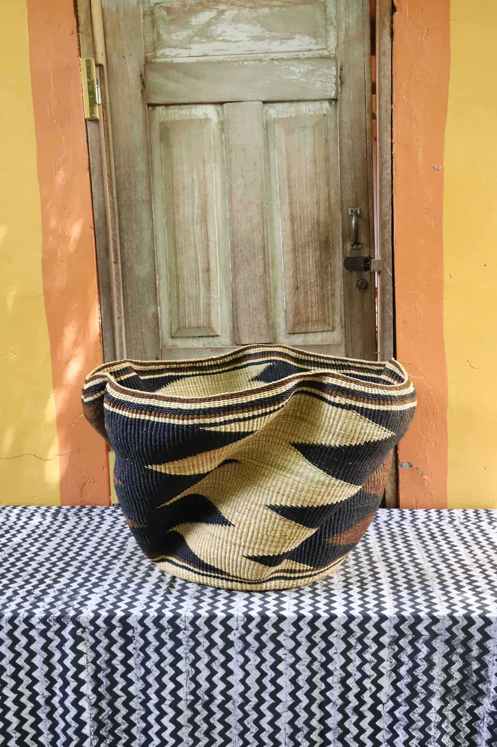 Giant Wave art basket in natural straw with bold black wave bands, on a black and white cloth before a wooden door.