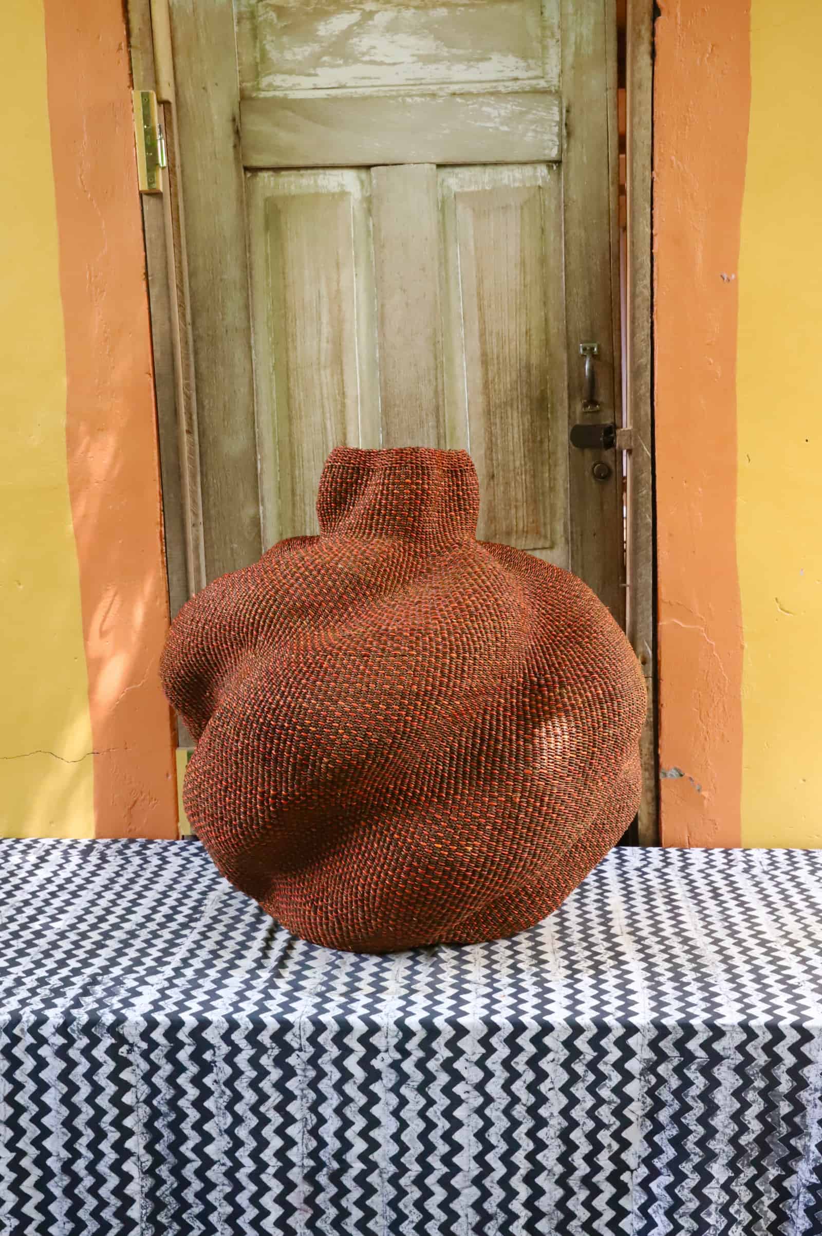 Large rounded woven basket in deep terracotta straw, on a black and white cloth against a wooden door