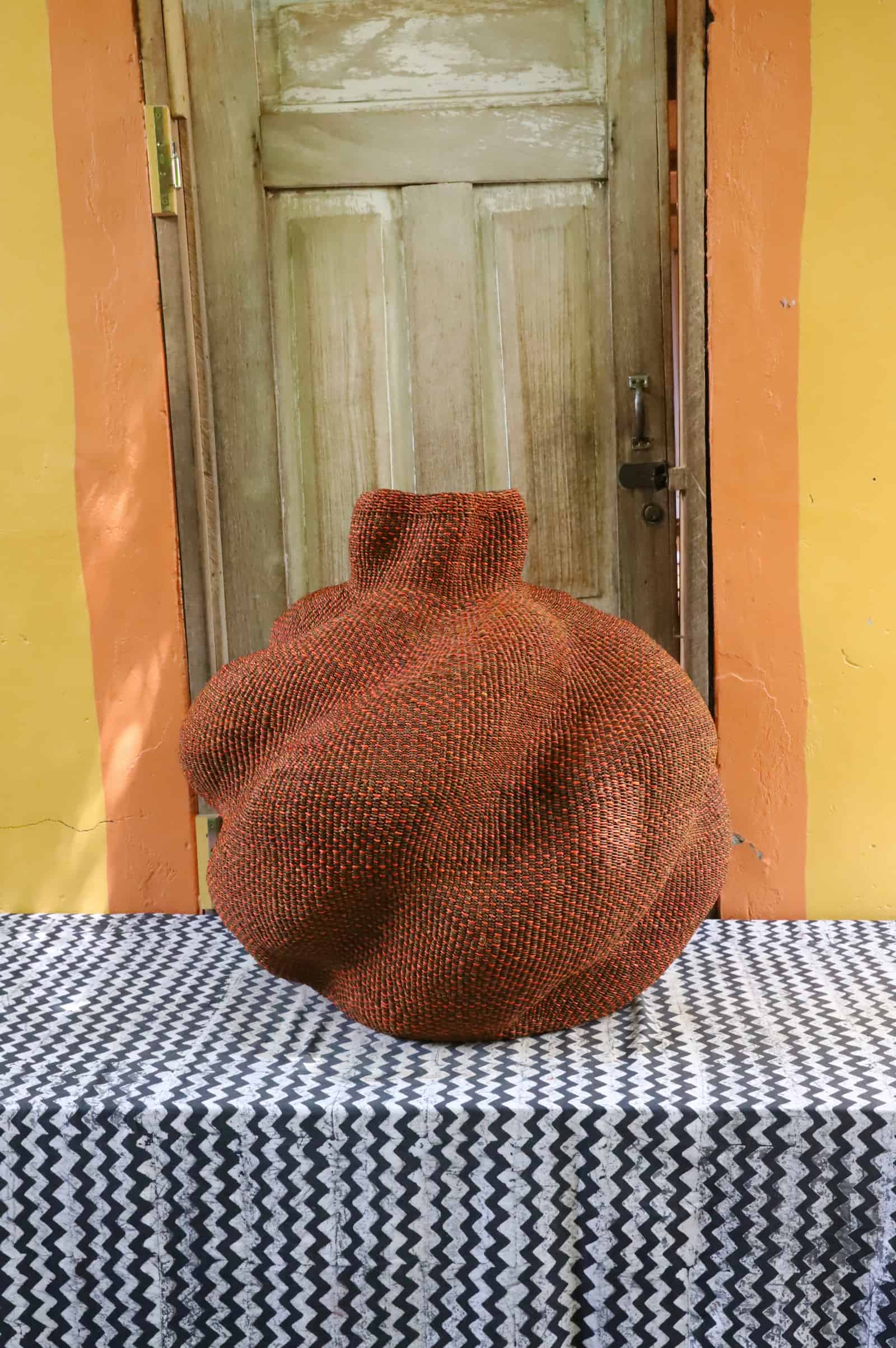 Large rounded woven basket in deep terracotta straw, different angle, on a black and white cloth, wooden door