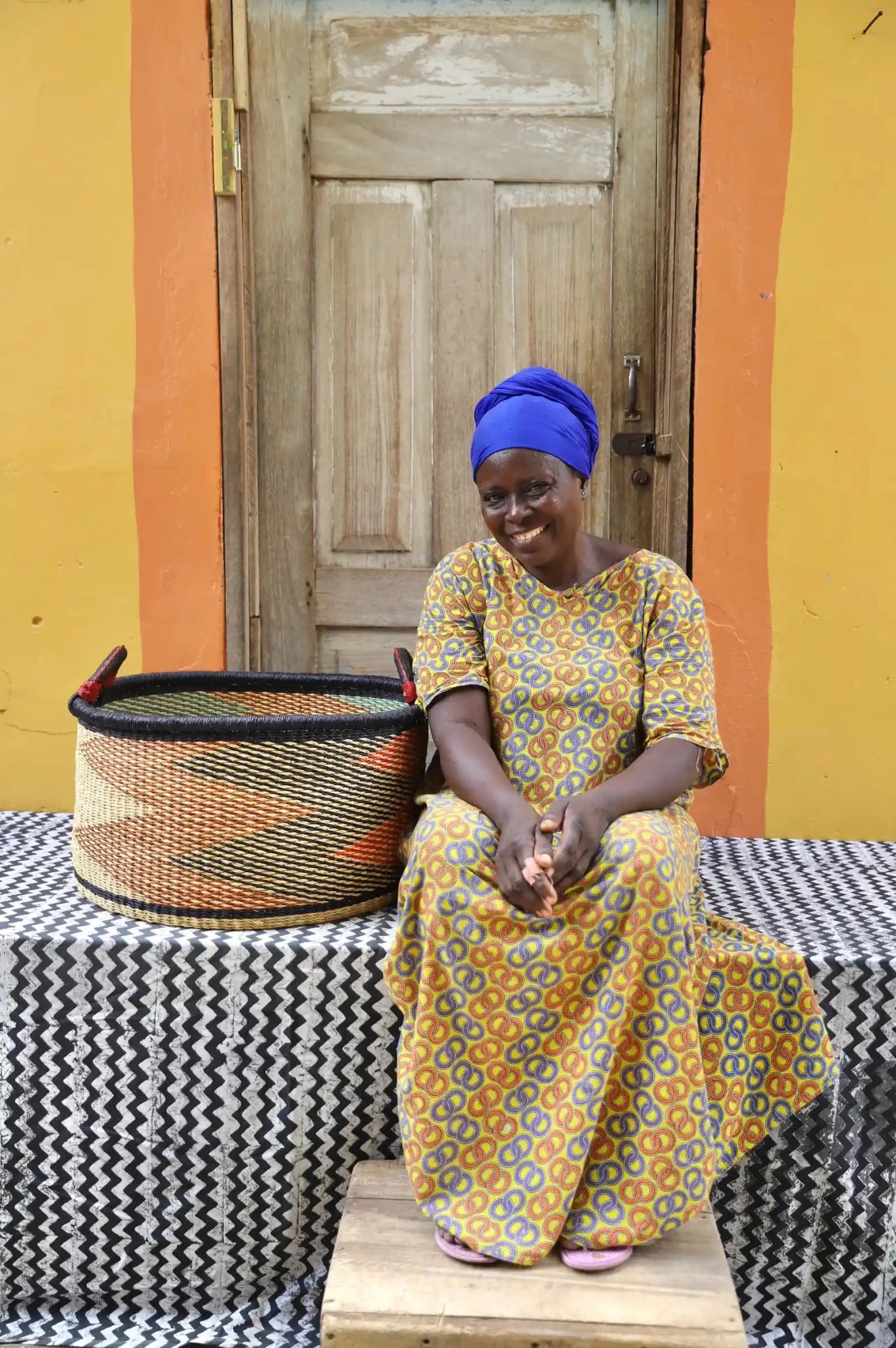 Woven Tub (Large) with woman weaver handwoven in Ghana sitting on black and white cloth and wooden door in background