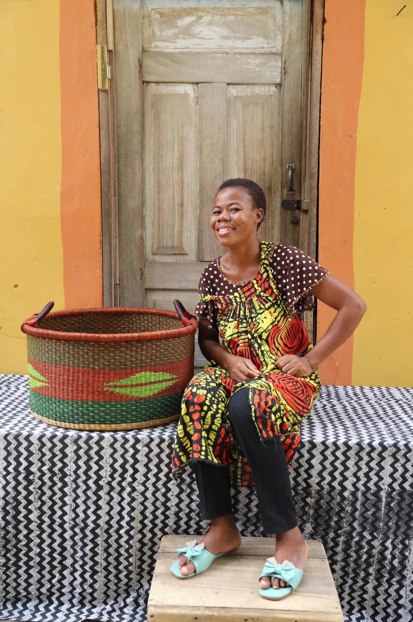 Woven Tub (Large) with woman weaver handwoven in Ghana sitting on black and white cloth and wooden door in background

