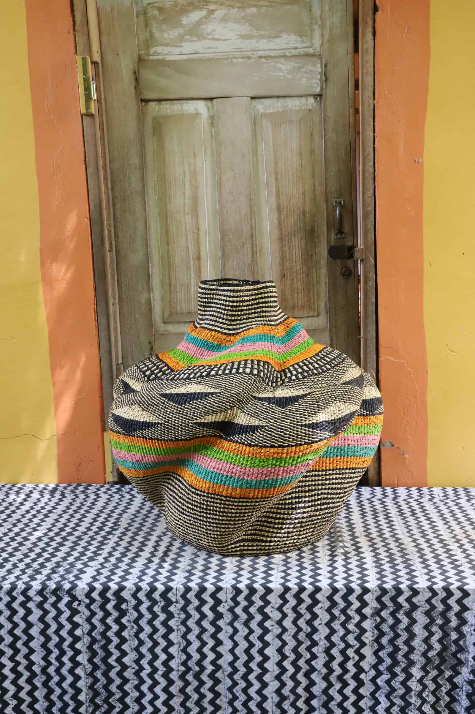 Rounded woven basket with bold orange, teal and pink bands on black pattern, on a black and white cloth against a wooden door