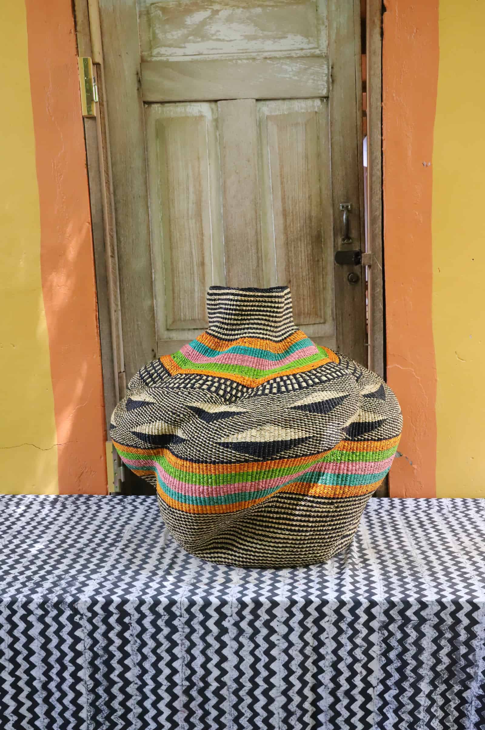 Rounded woven basket with orange, teal and pink bands on black pattern, rear view on a black and white cloth, wooden door