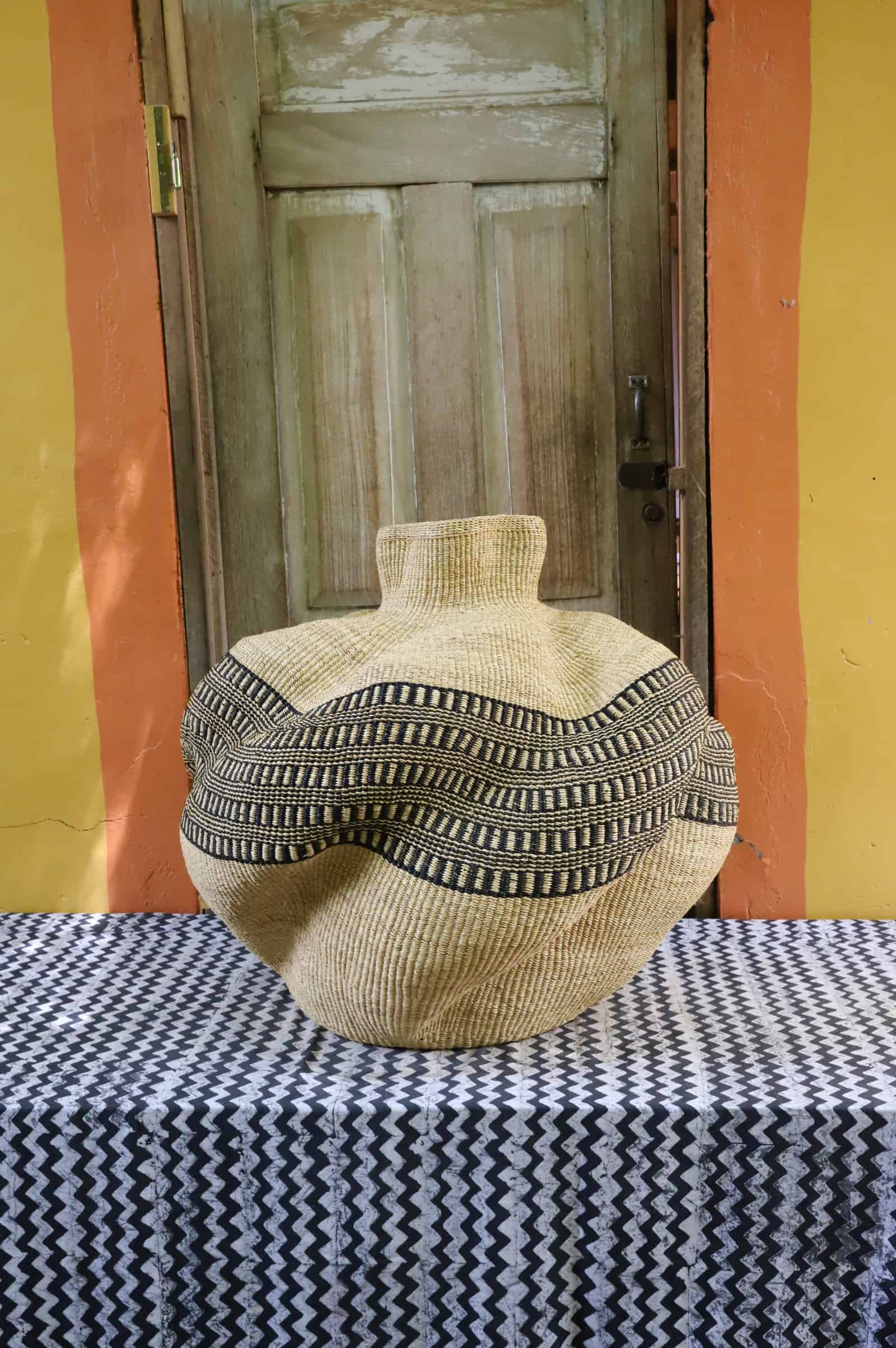 Bulbous woven basket in natural straw with a black and cream geometric band, on a black and white cloth against a wooden door