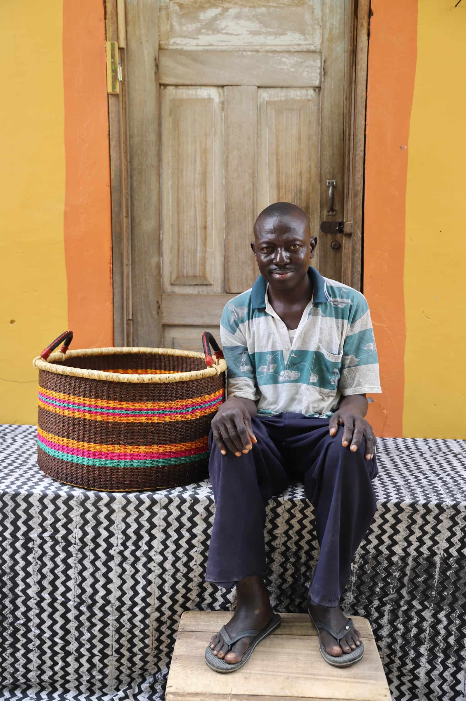Woven Tub (Large) with man weaver handwoven in Ghana sitting on black and white cloth and wooden door in background

