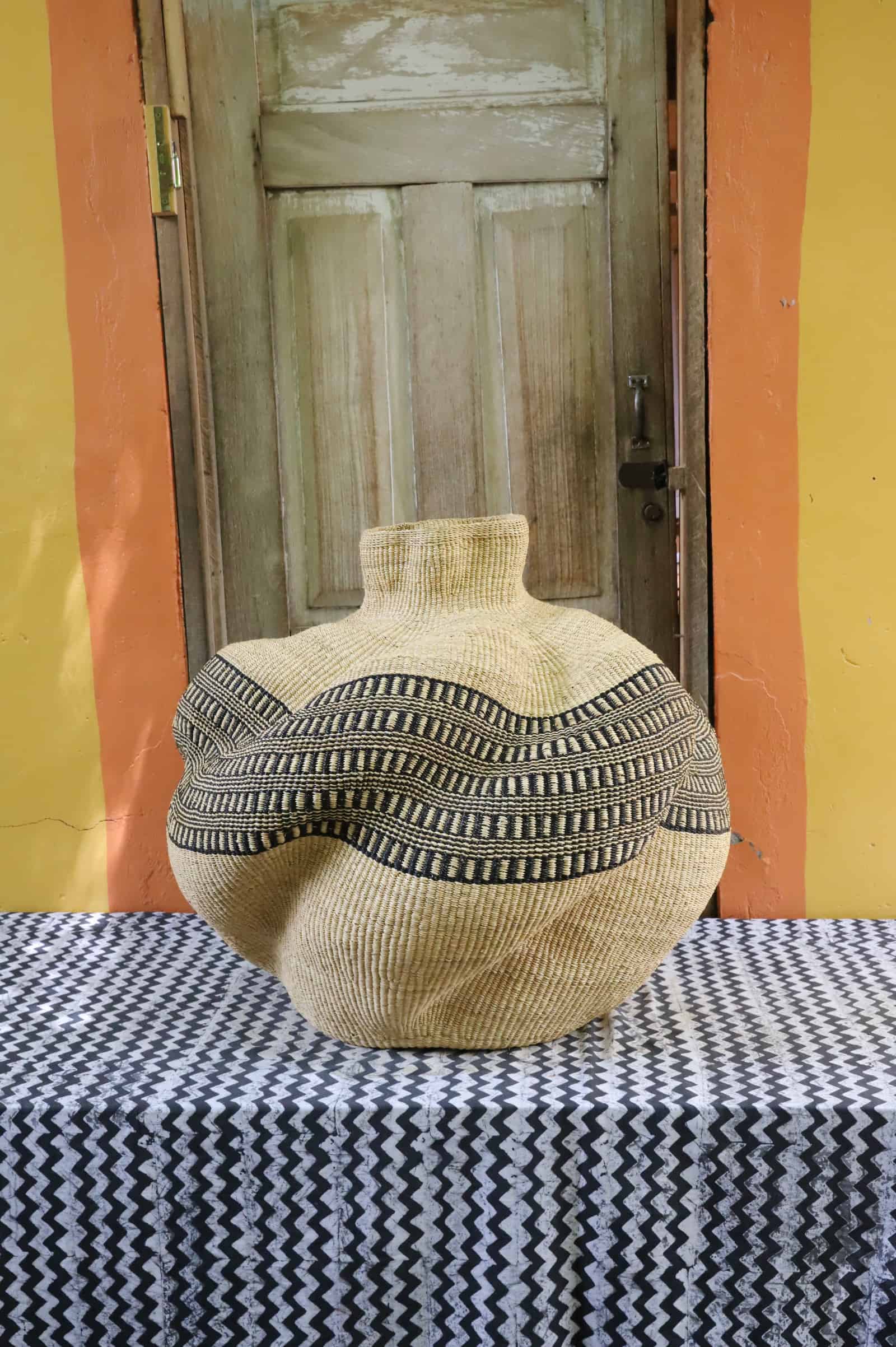 Bulbous woven basket in natural straw with black and cream geometric band, side view on a black and white cloth, wooden door