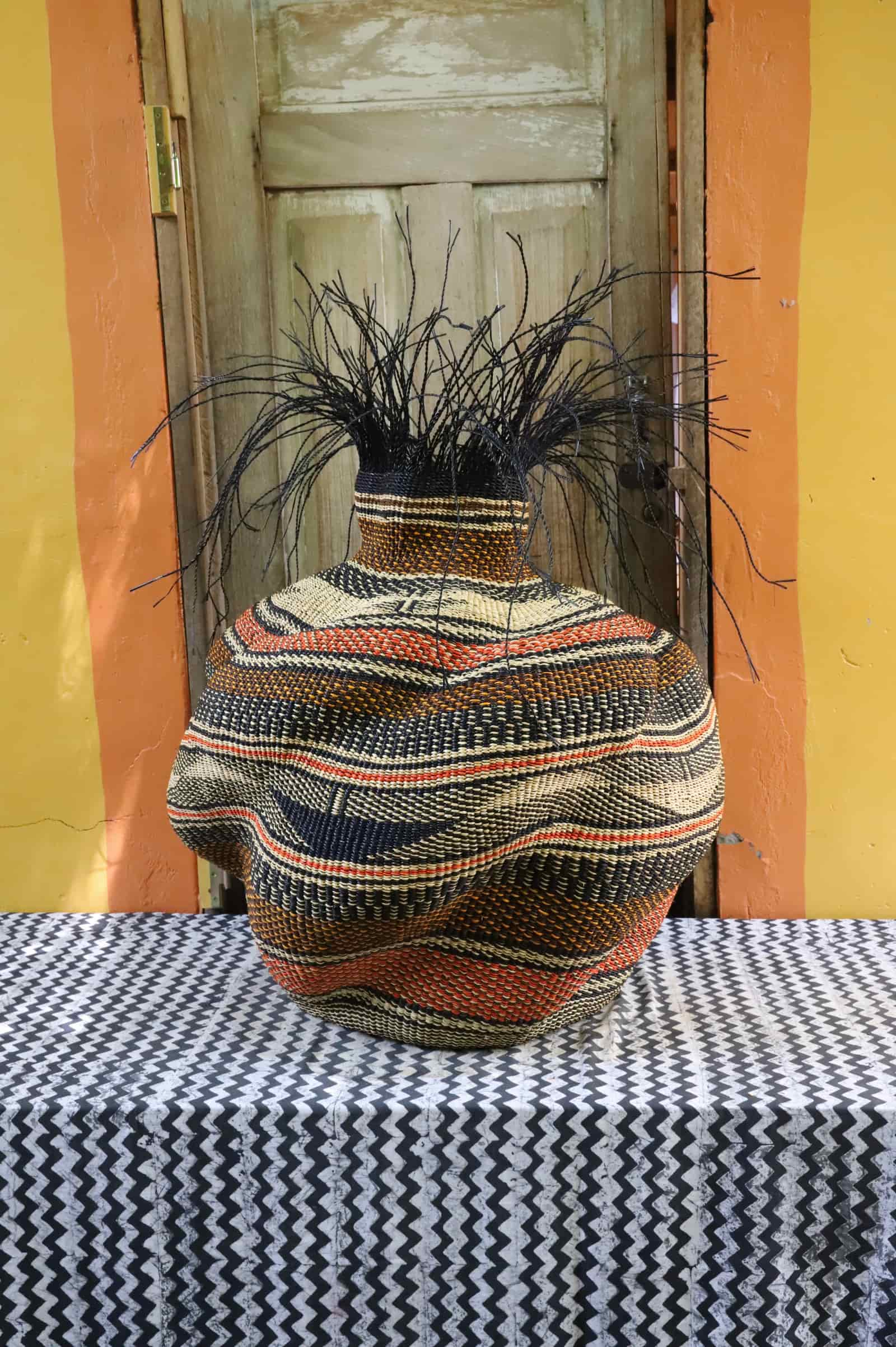 Large Tua-Tia baobab art basket in dark and terracotta stripes with black straw branching, on chevron cloth against a wooden door.