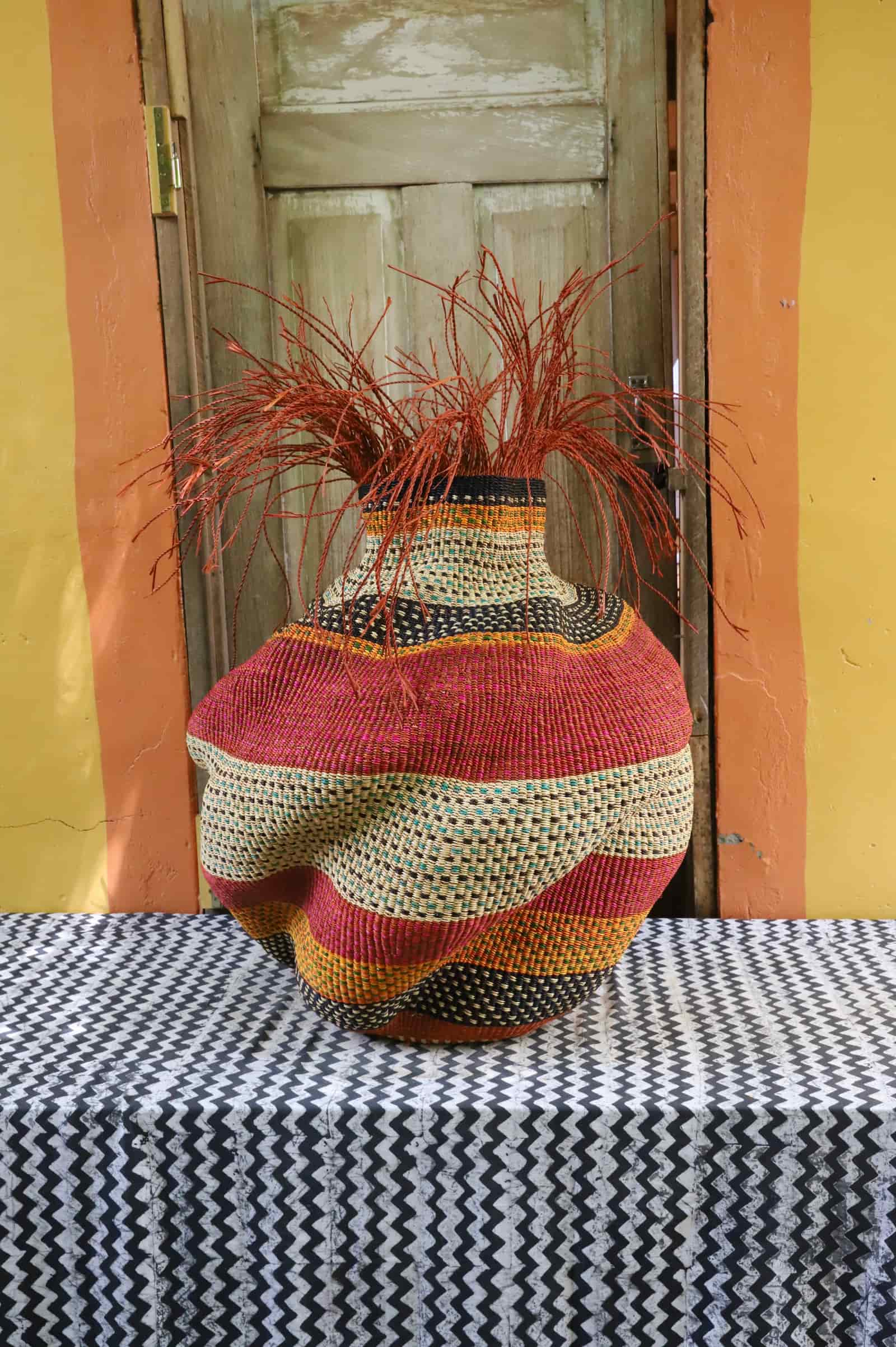 Large Tua-Tia baobab art basket in red and natural stripes with red straw, side view on chevron cloth against a wooden door.